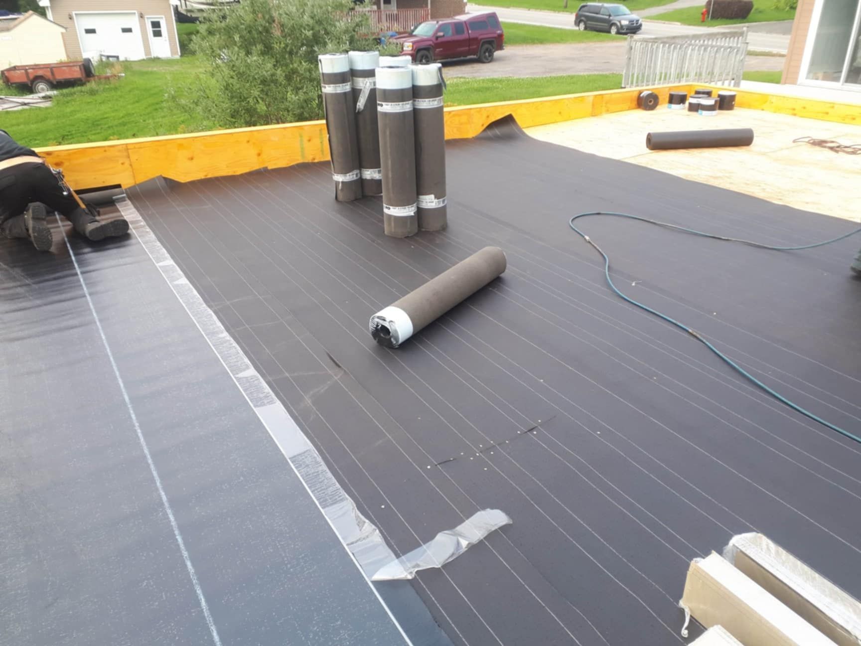 Matériau de toiture noir en cours d'installation sur un toit plat, rouleaux de matériau présents, cadre extérieur.