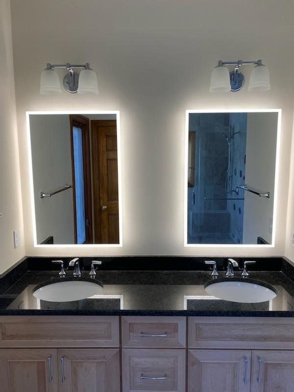 Two illuminated vanity mirrors above matching sinks and a black countertop.
