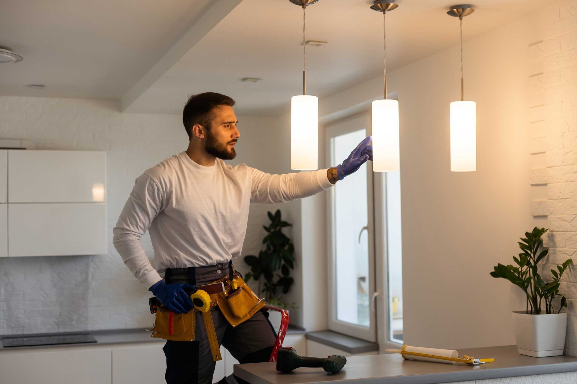 Electrician on a stepladder installing light fixtures in a kitchen, wearing a tool belt.