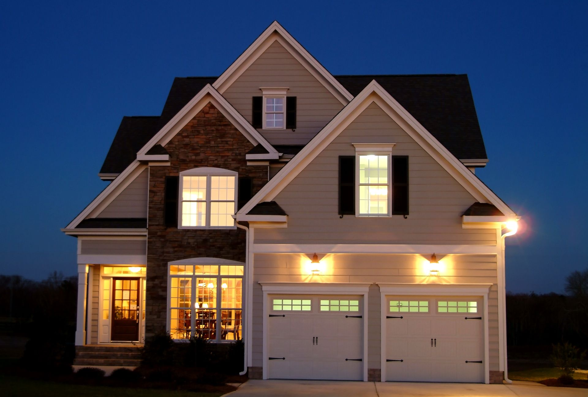 Two-story house at dusk, lit interior lights, stone and tan exterior, two-car garage.