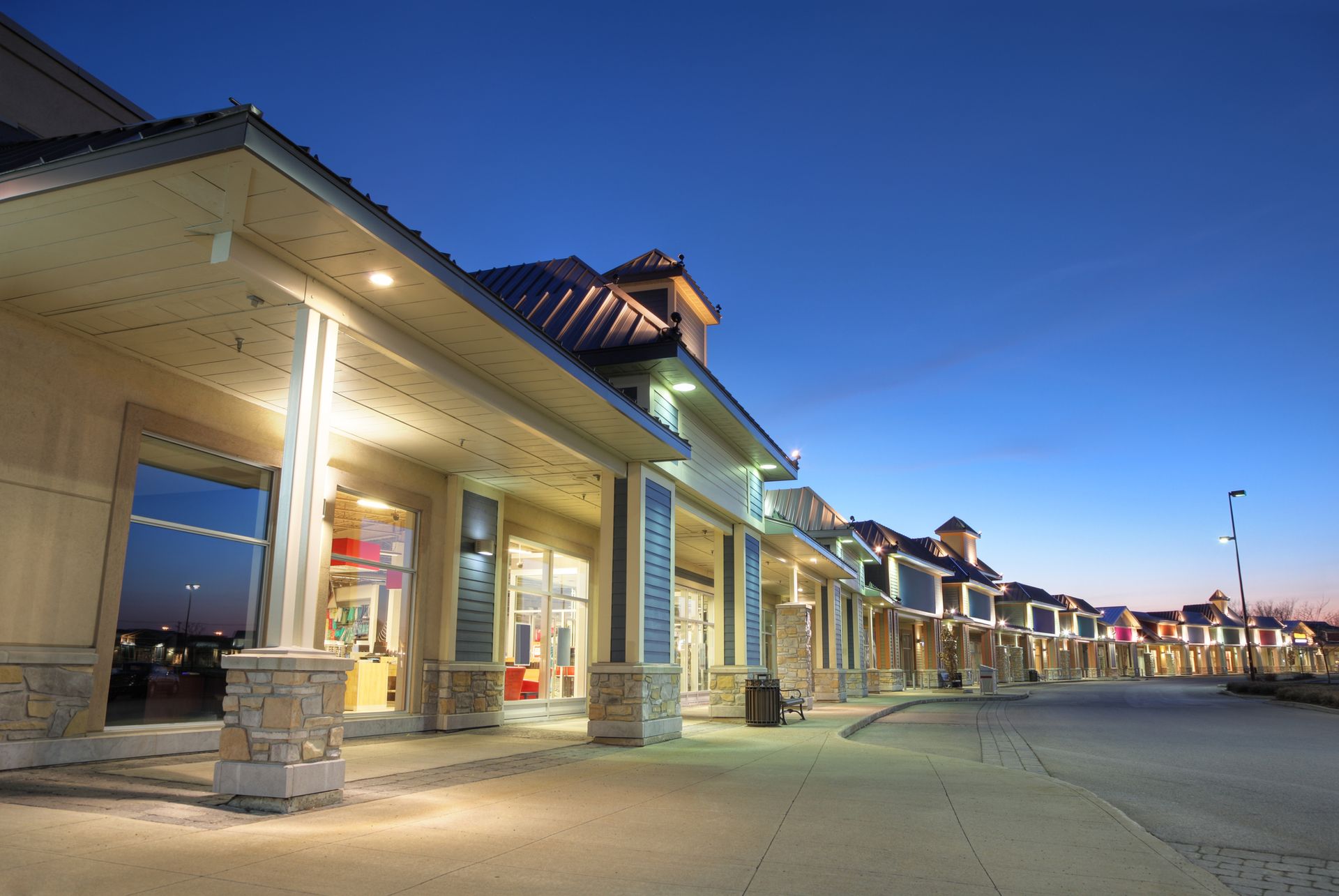 Shops in a strip mall at dusk; illuminated storefronts and columns with a blue sky.