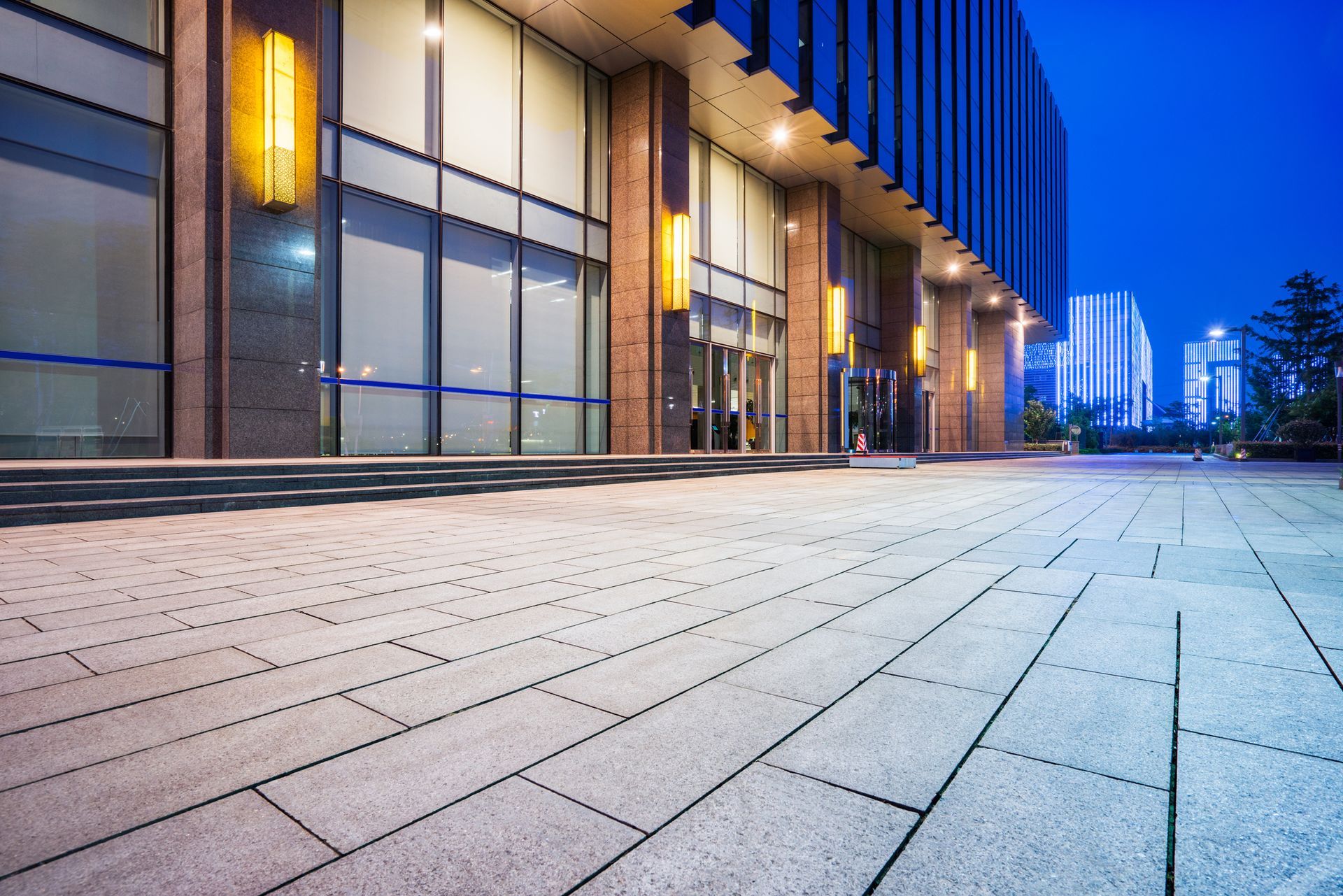 Building exterior at dusk, large glass windows, brick pavers, illuminated lights.
