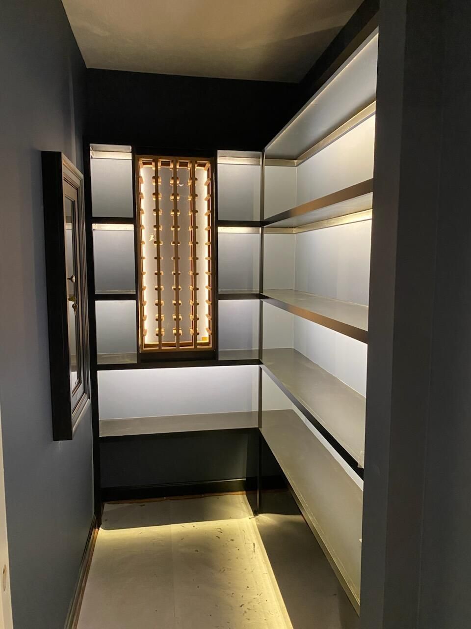 Walk-in closet with illuminated shelves and a wine rack, featuring white, gold, and dark gray accents.