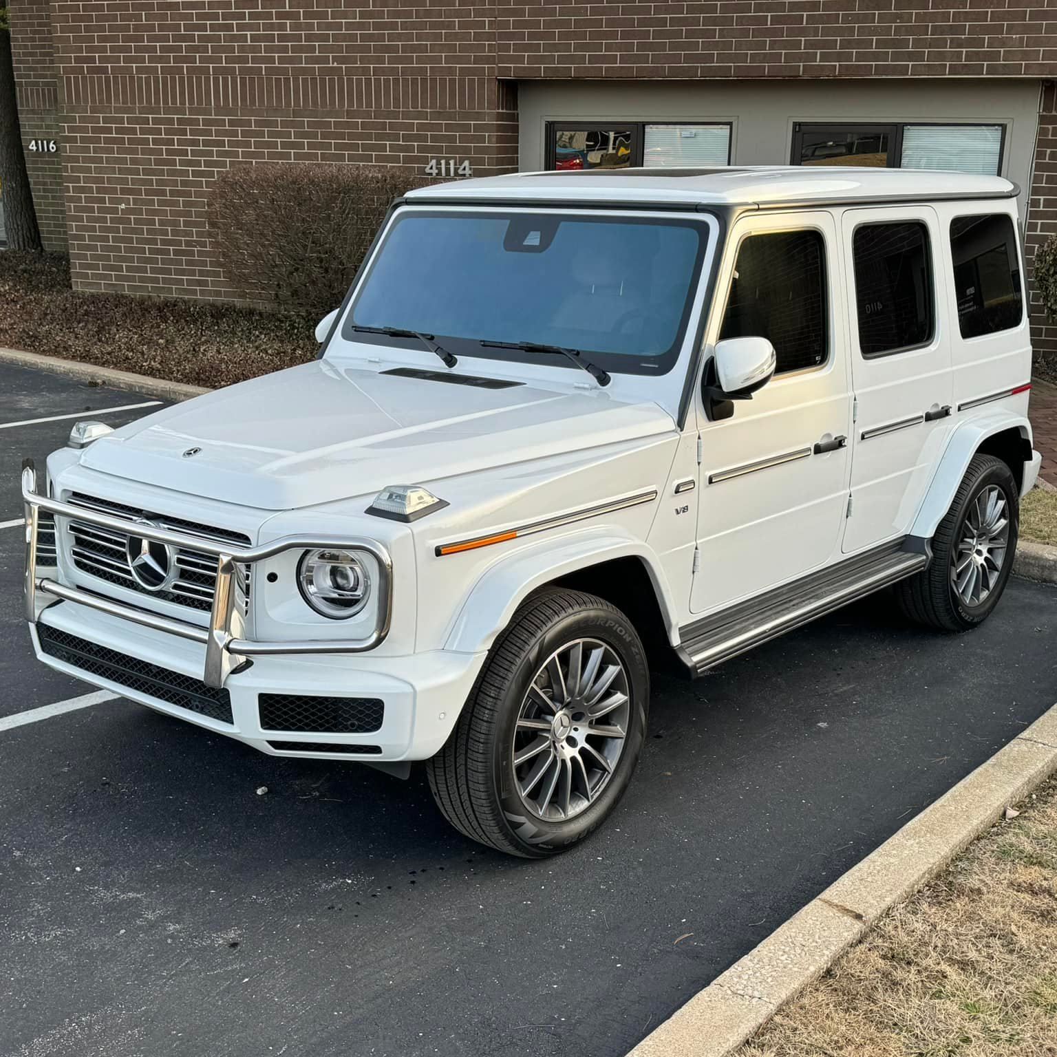 A white mercedes benz g class is parked in a parking lot in front of a brick building.