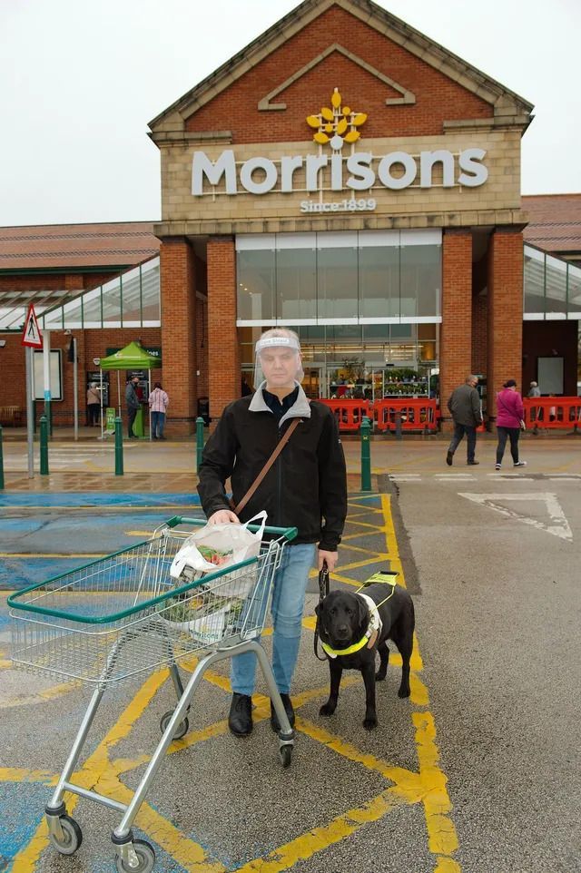 A man wearing a black jacket, blue jeans, and a transparent face shield stands outside a Morrisons.