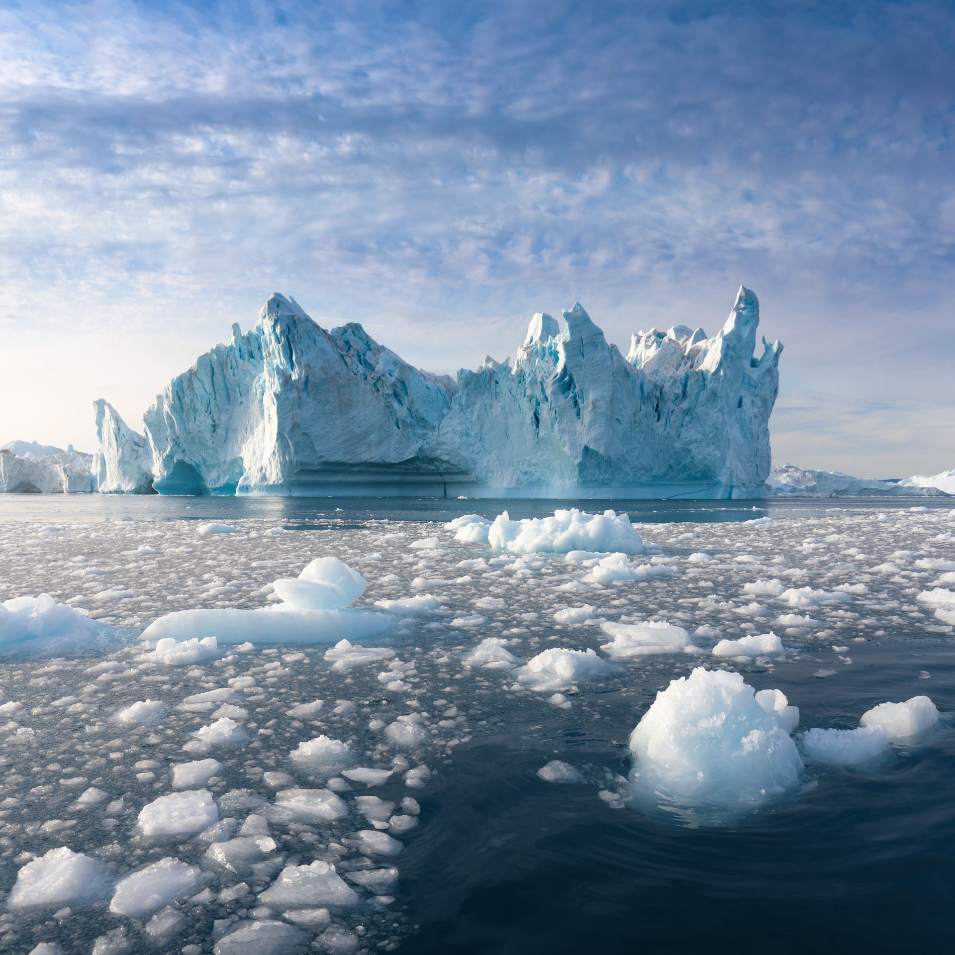 A glacier juts against a cloudy sky as if a crown, small sharp segments disintegrating away and crumbling into the surrounding sea.