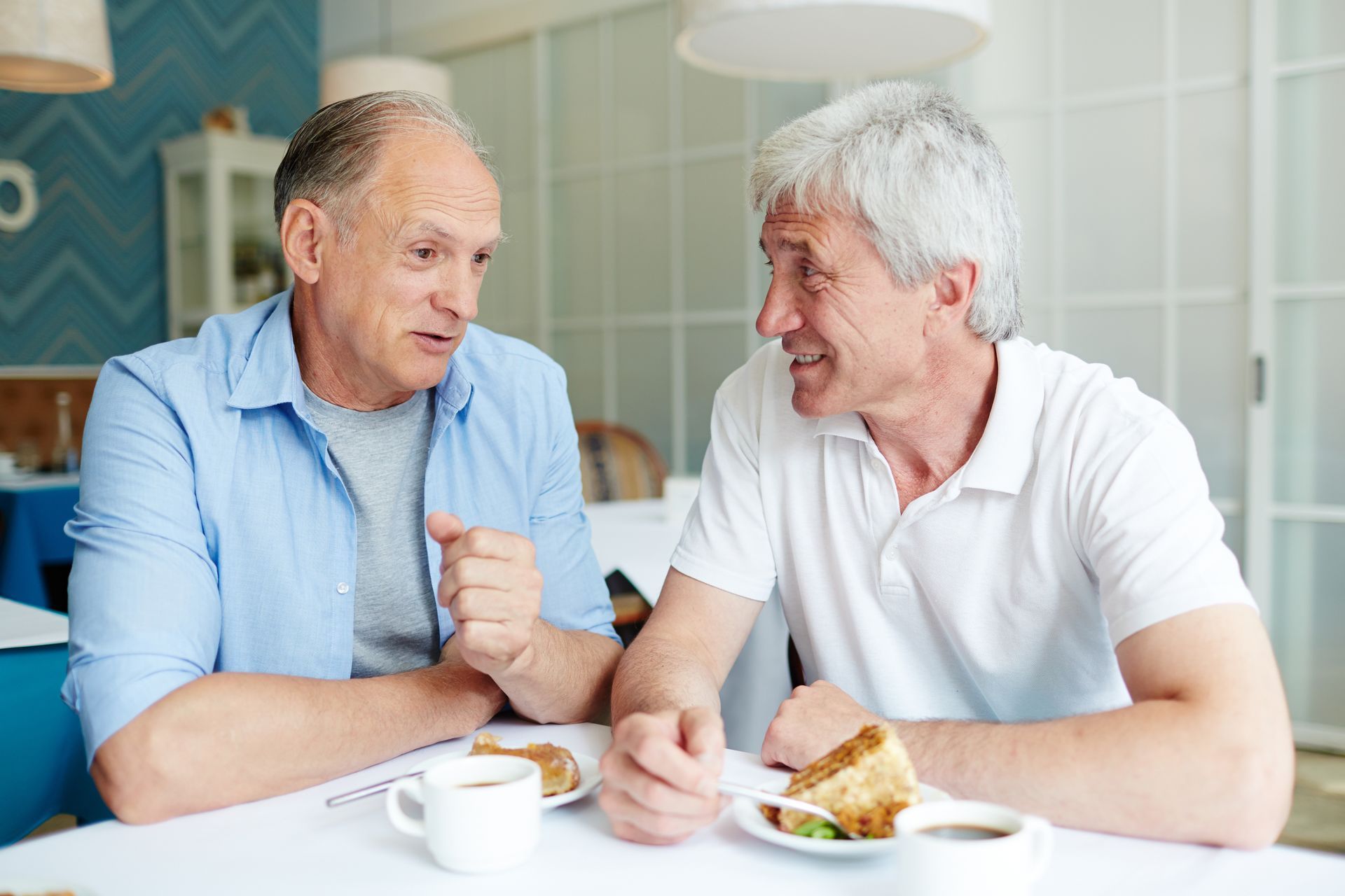 (Communication is Key) Two mature men laugh and chat together over cake in a small, cosy café.