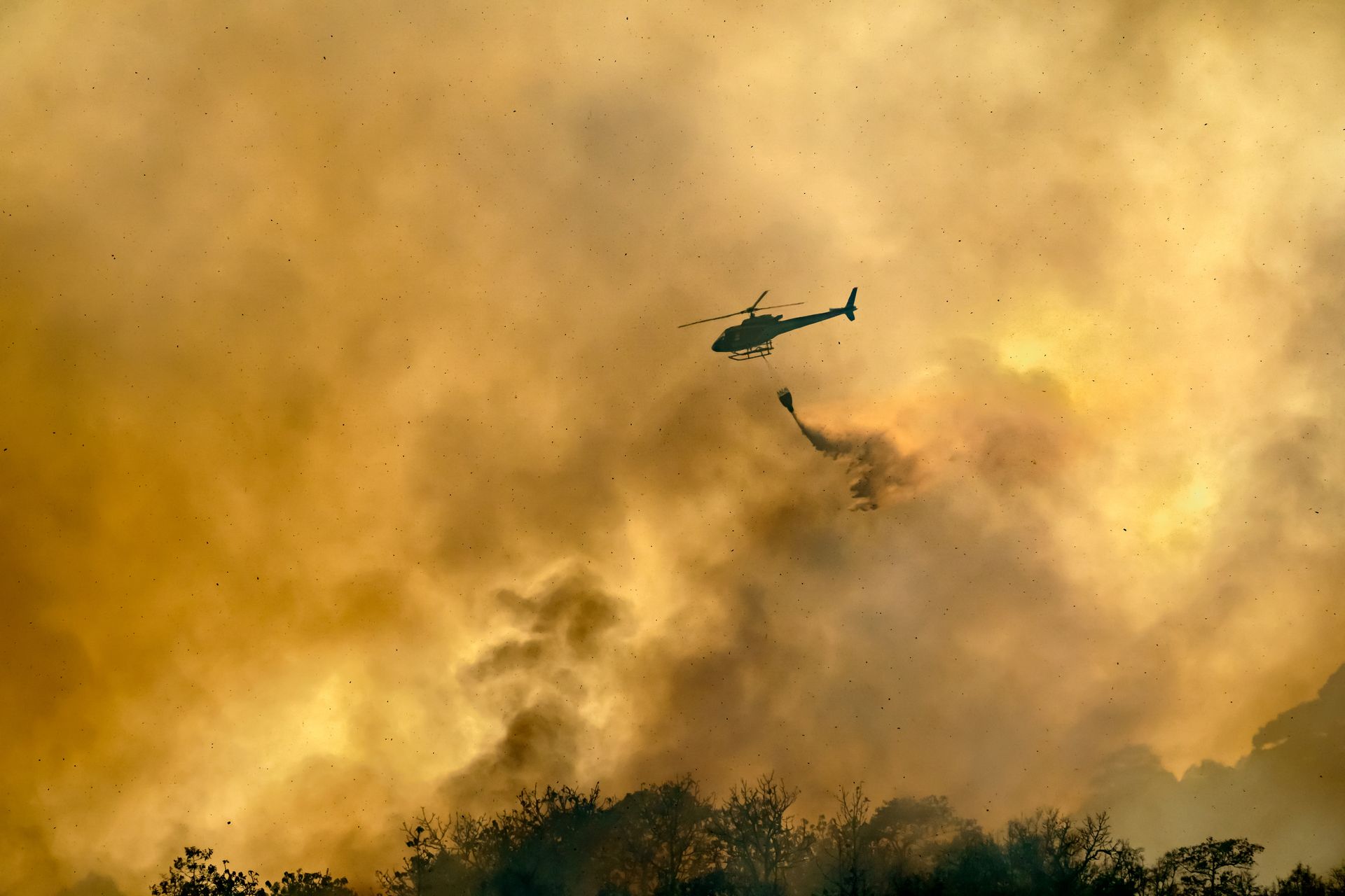 A helicopter hovers over a raging wildfire, releasing a burst of water that seems to fade into the towering smoke.