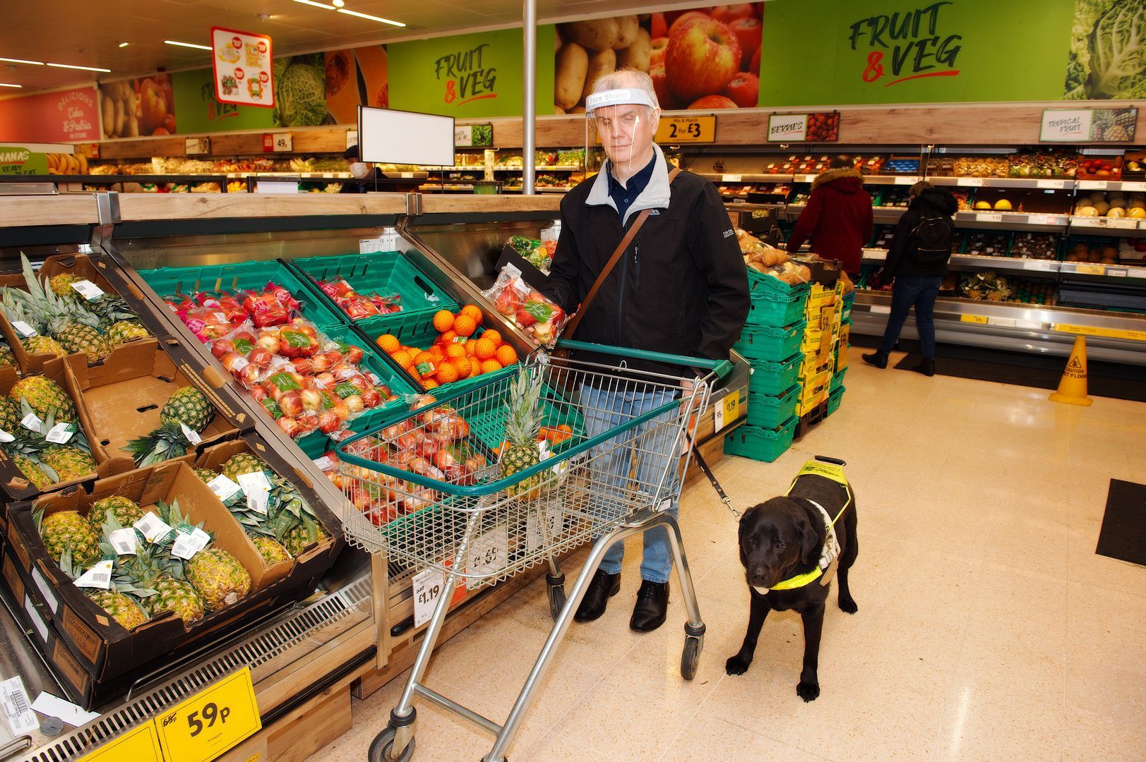 Philip pushes a shopping trolley down the fruit aisle, selecting a pineapple. His guide dog waits by his side.