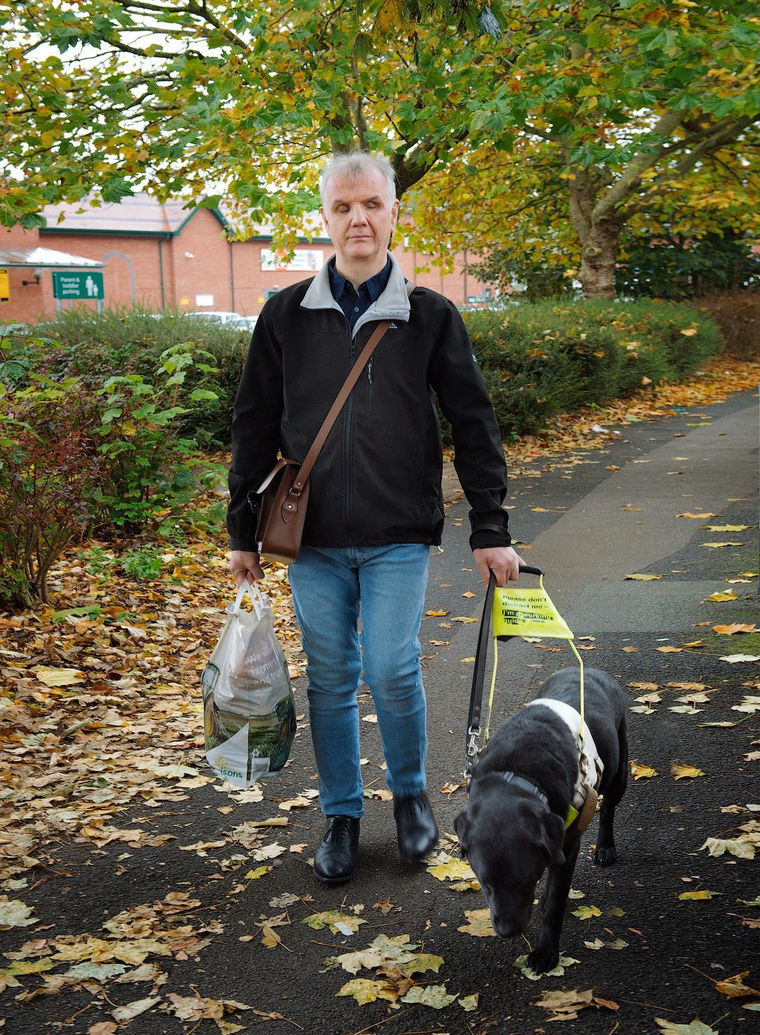 Philip walks down a leaf-strewn path with his guide dog, shopping bag in hand.