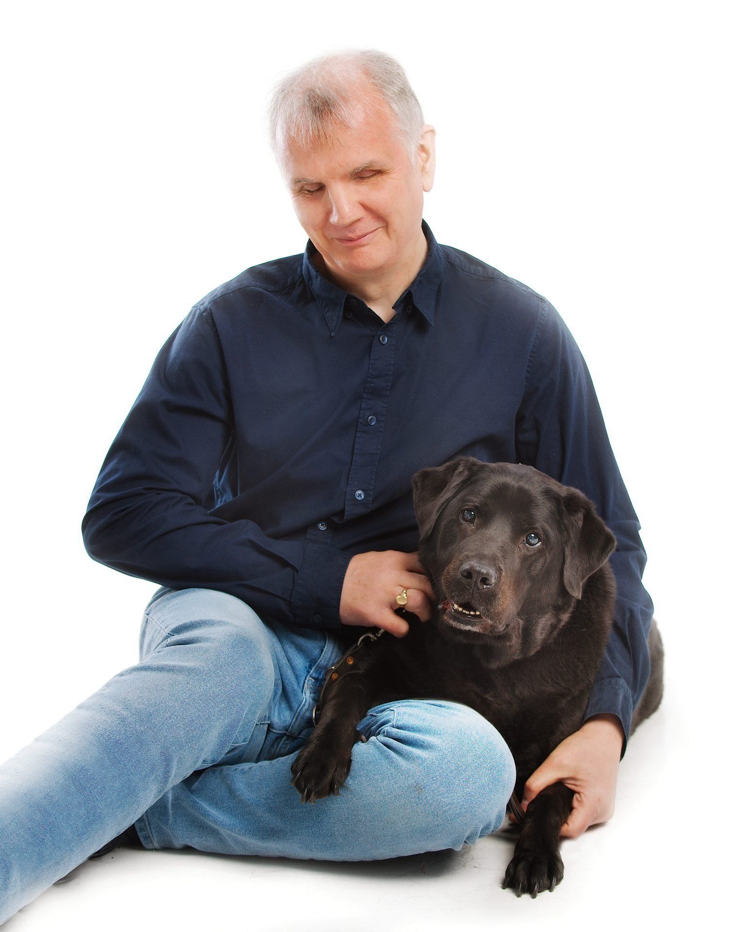 Informal pose of Philip Anderson sitting on the floor with one arm around his black guide dog, Quintana, who has one paw resting on his knee
