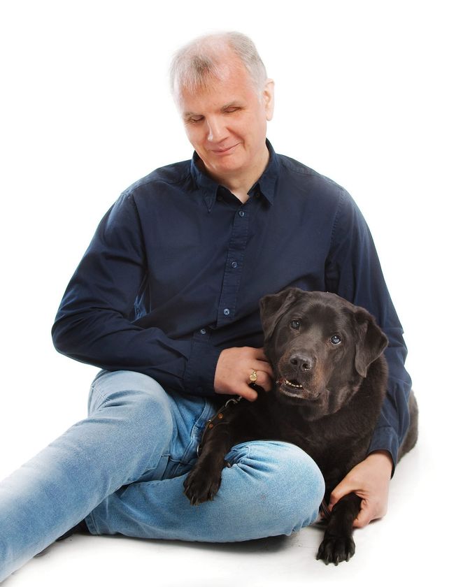 Informal pose of Philip Anderson sitting on the floor with one arm around his former black guide dog, Quintana, who has one paw resting on his knee