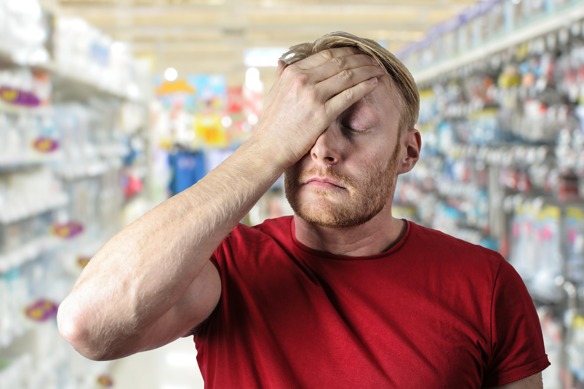 A man stands in a supermarket, rubbing at his face in frustration.