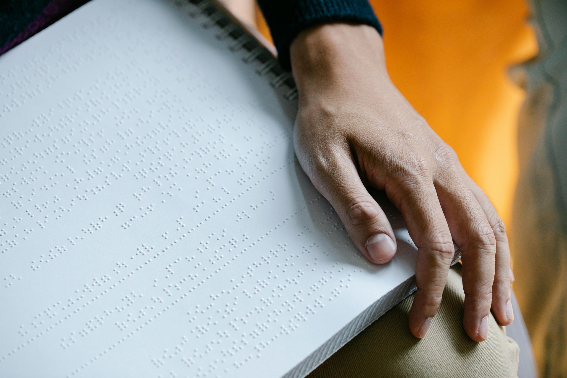 A close-up of a person's hand resting on the edge of an open Braille book. The hand is positioned over the right-hand page, which is covered in raised Braille dots arranged in lines. The person is wearing a dark long-sleeved top, and the book is spiral-bound. The background includes a soft orange hue and part of the person's khaki trousers.
