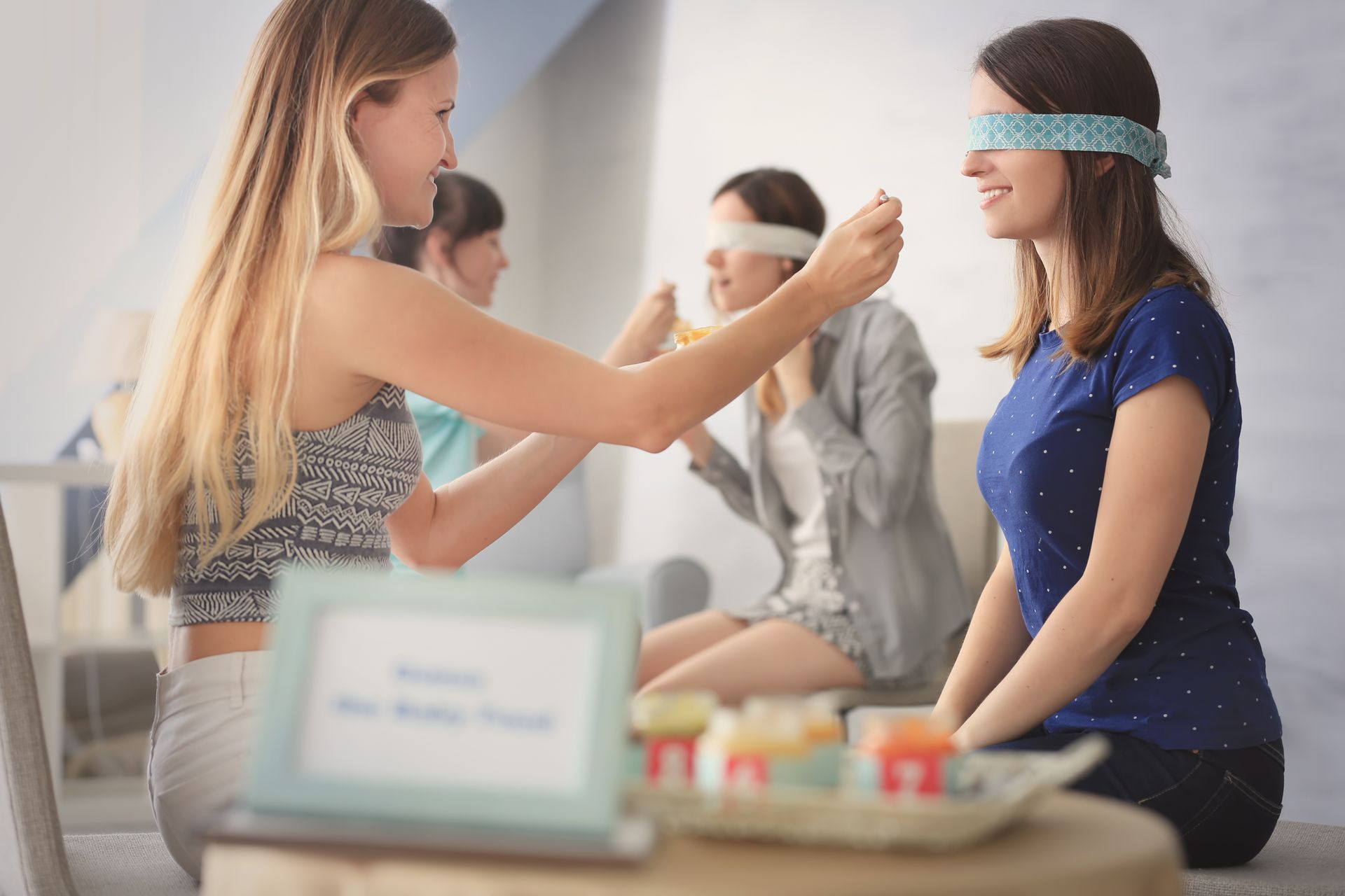 In the foreground, a woman in a blindfold is fed by another woman. In the background, another woman in a blindfold attempts to feed herself.