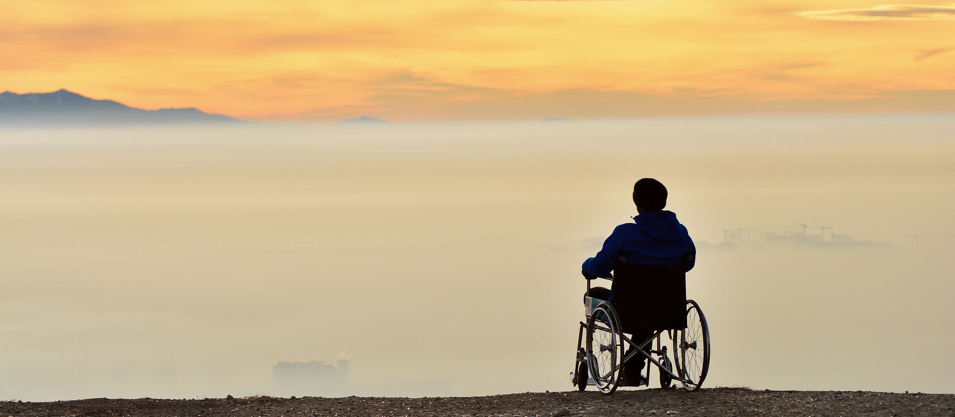 A man in a wheelchair stares out at the horizon from atop a mountain as the sun rises above a misty valley.