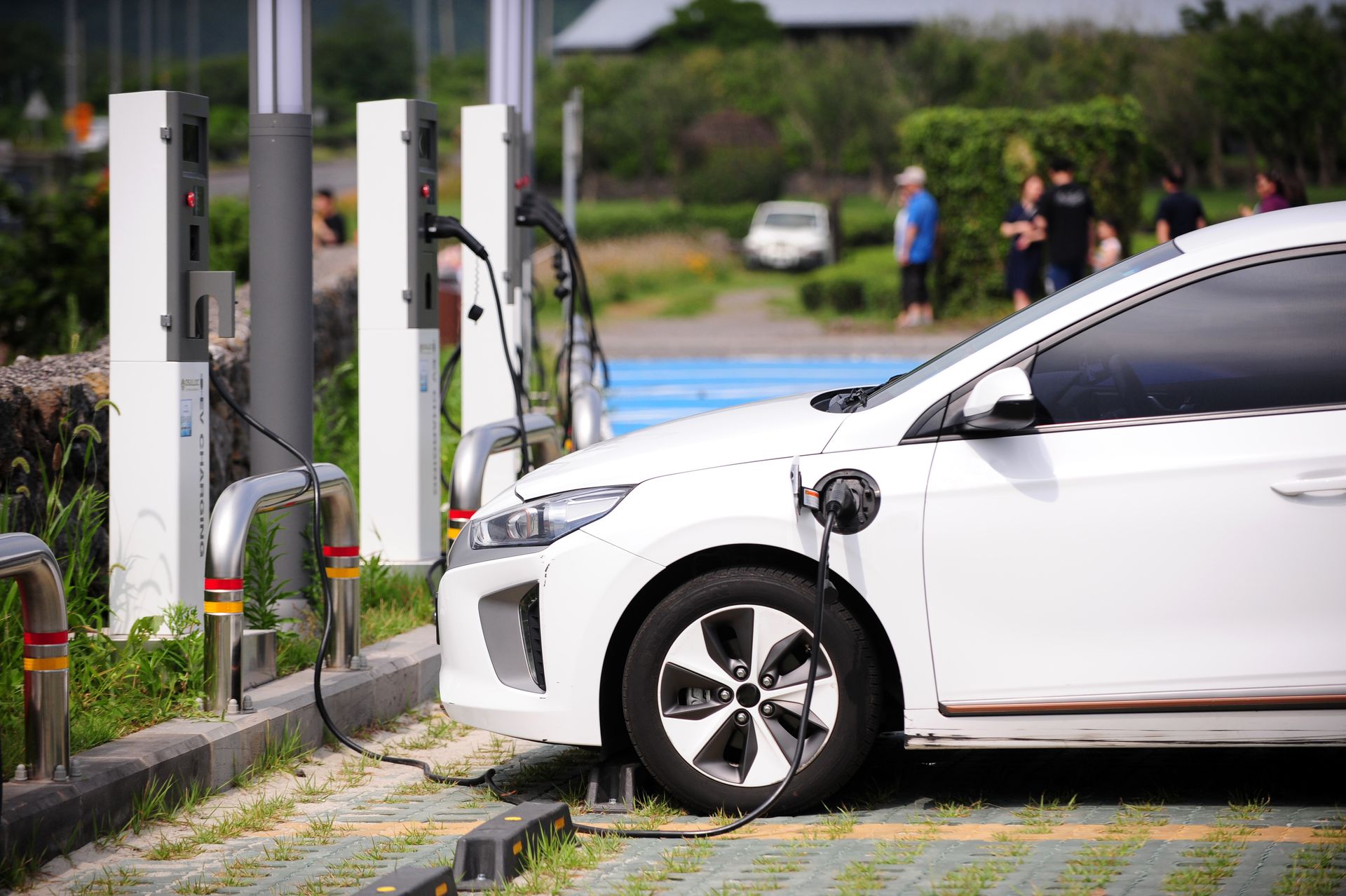 Photo of a white car charging at an EV charge station. The ground is uneven, with tufts of grass and a raised pavement.