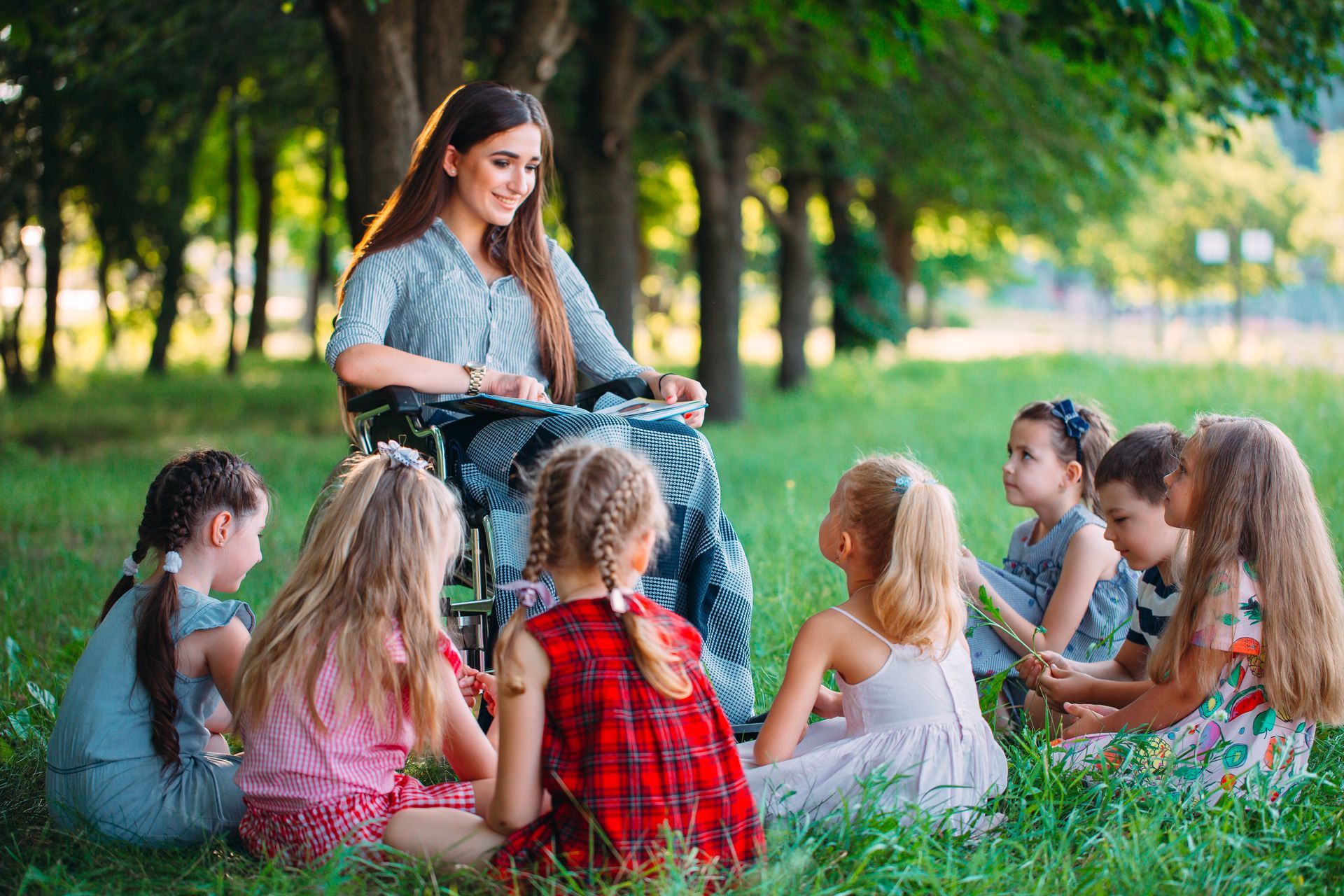 A young and beautiful female teacher in a wheelchair conducts a class outside with a group of smiling, attentive children.