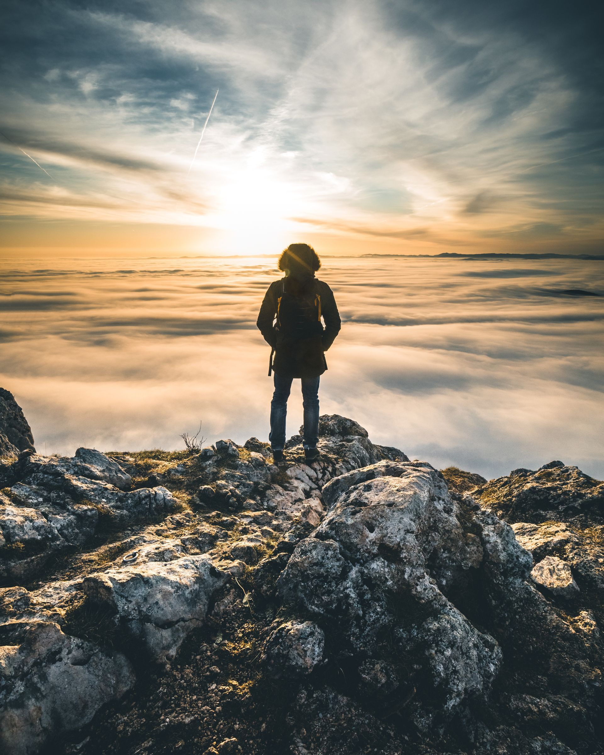 A hiker stands at the peak of a snowy mountain, gazing into a blazing sunset and a blanket of clouds below. 