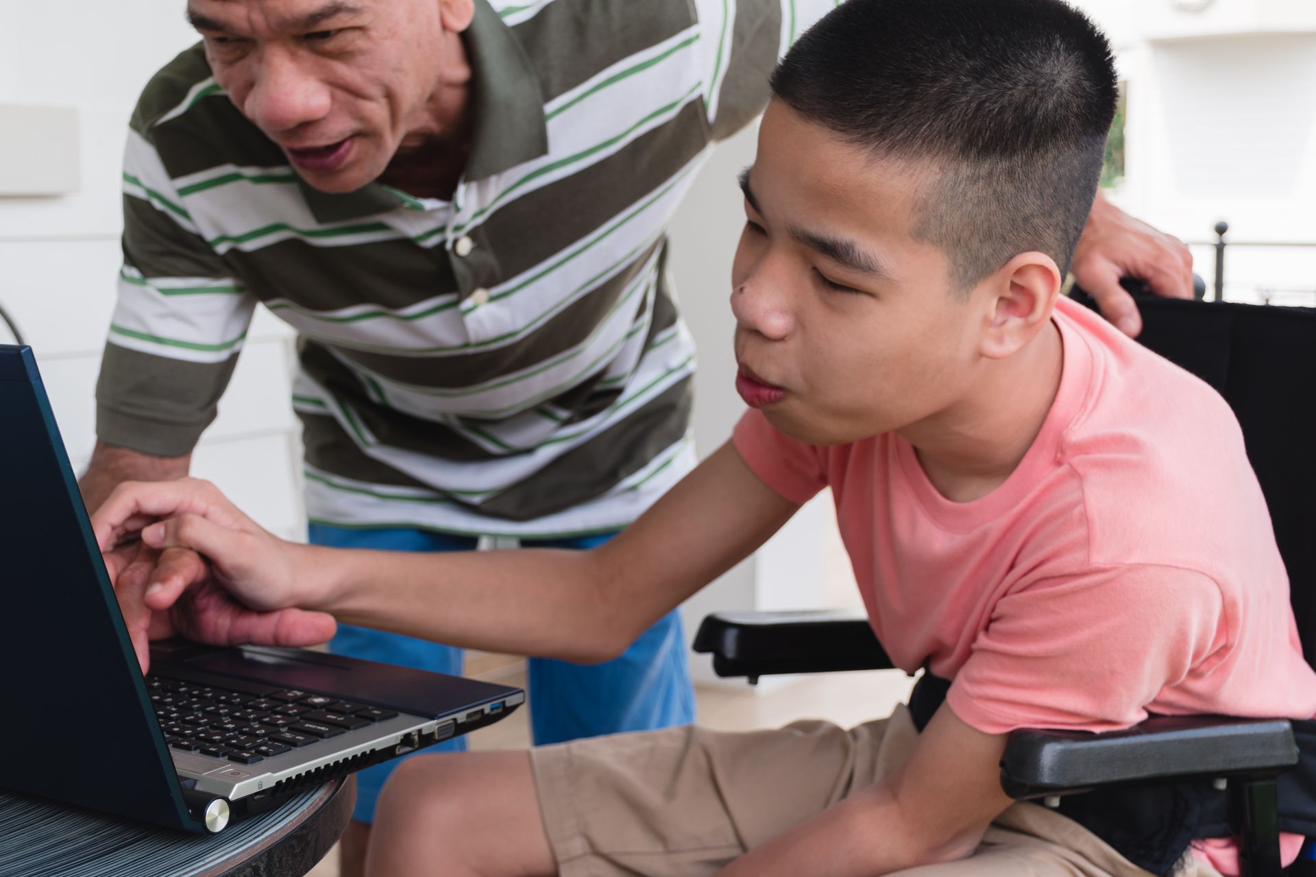 A young boy with a cognitive disability leans over a computer with an older male, pointing towards the screen