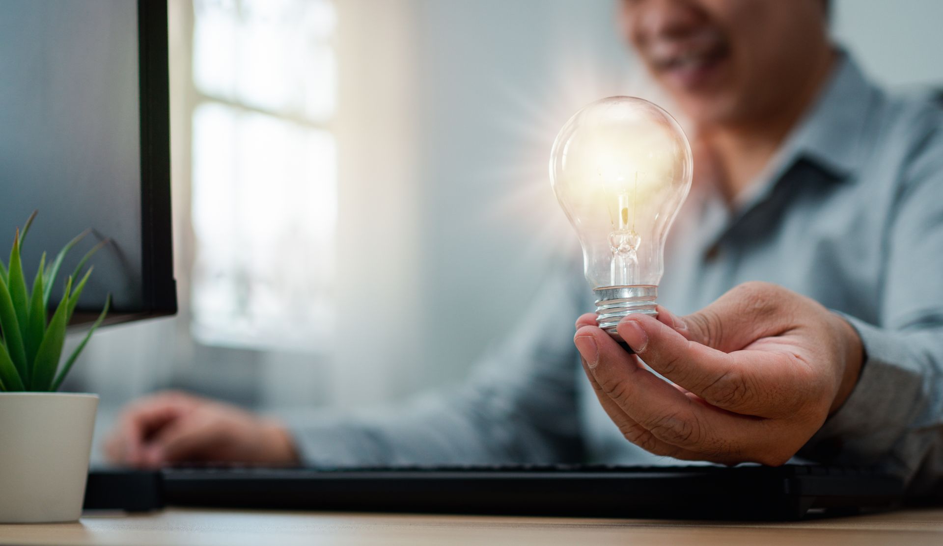 A photograph of person sat at a desk holding a light-bulb. The bulb is shining brightly.