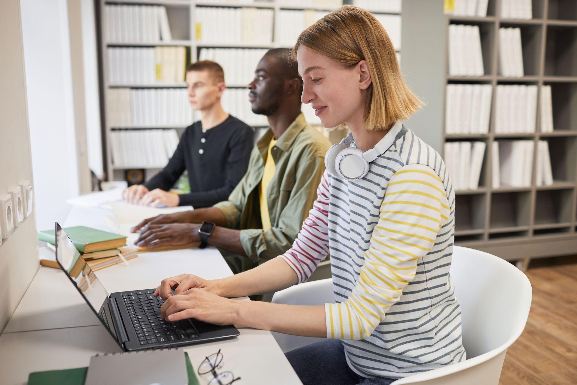 A young woman studies in the library using her laptop. Studying alongside her are two young men are researching using Braille.