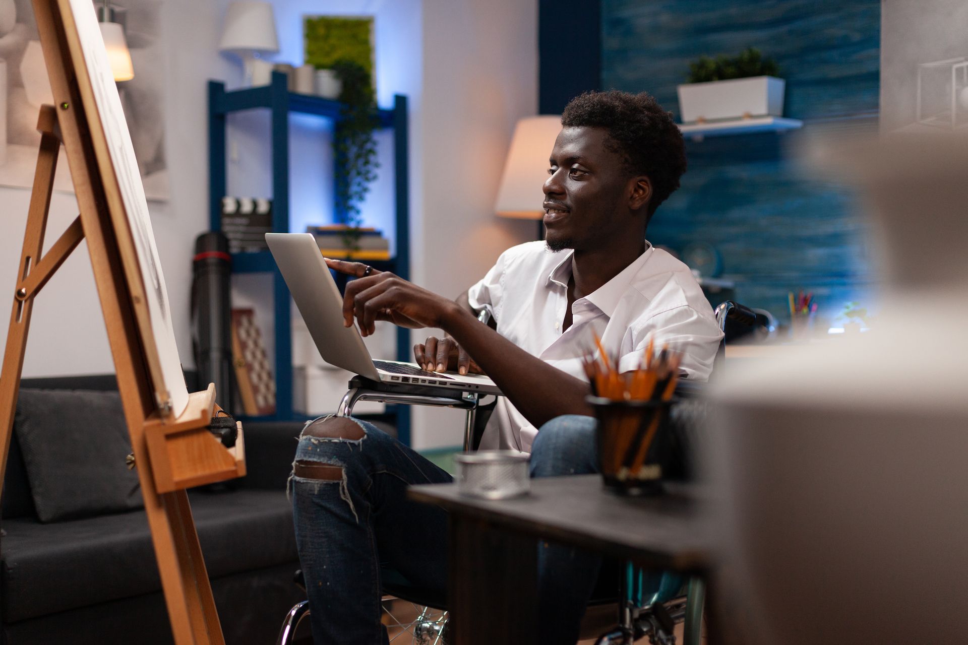 A young illustrator in wheelchair uses a laptop while looking at an easel. 