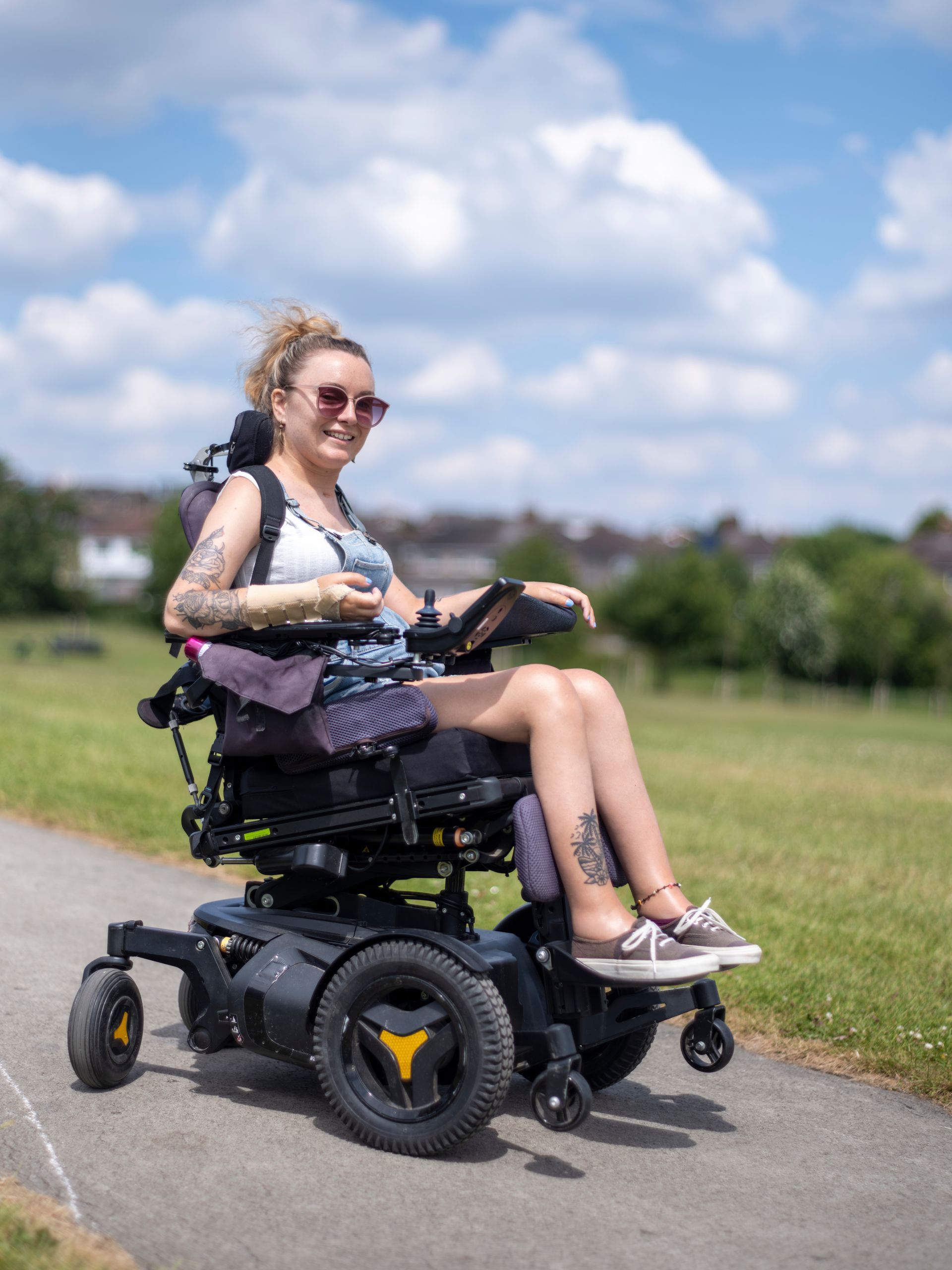 A smiling woman in a motorised wheelchair enjoys a bright and sunny day in the park.