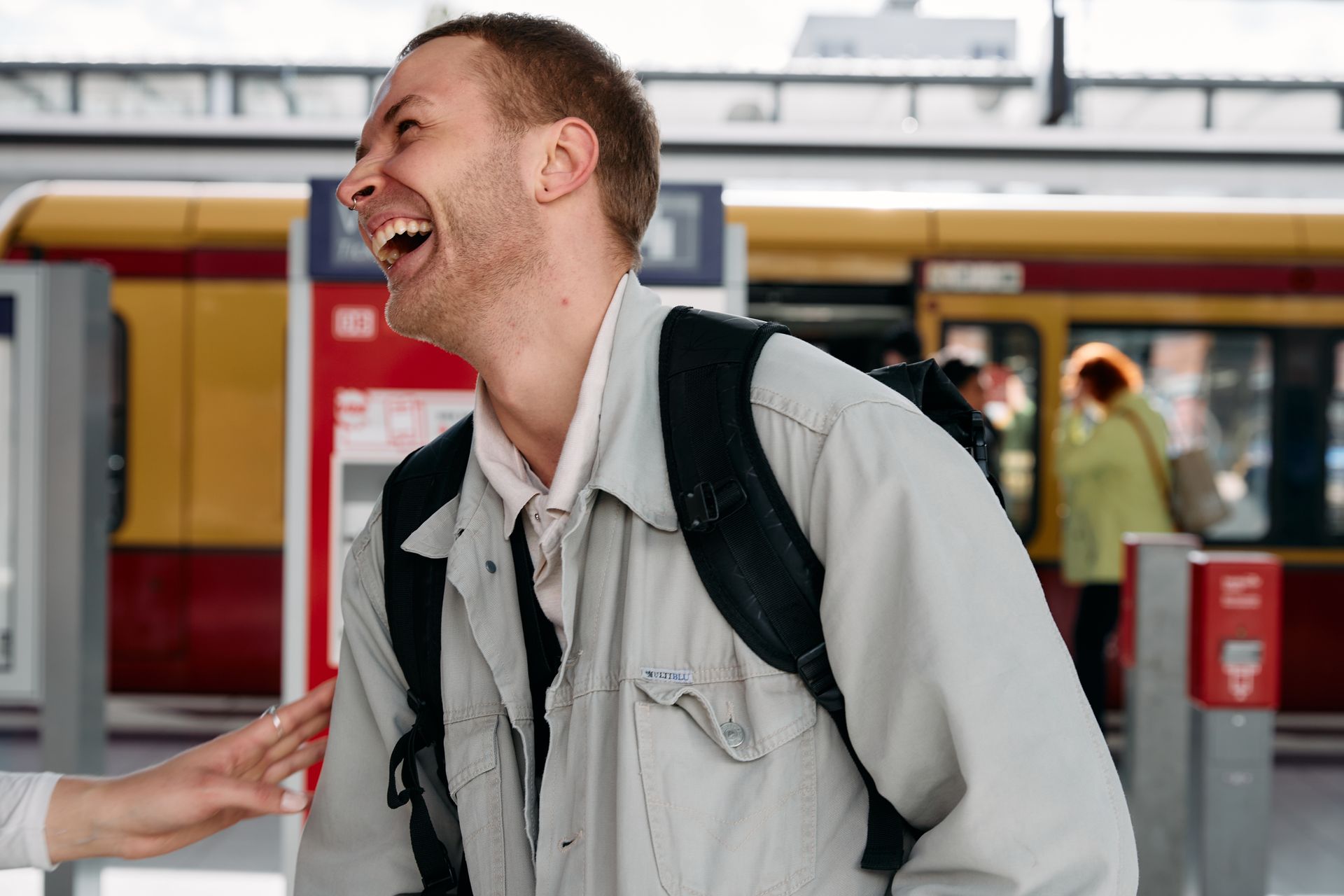 A young man stood in front of a busy train platform laughs as he pays at a ticket booth.