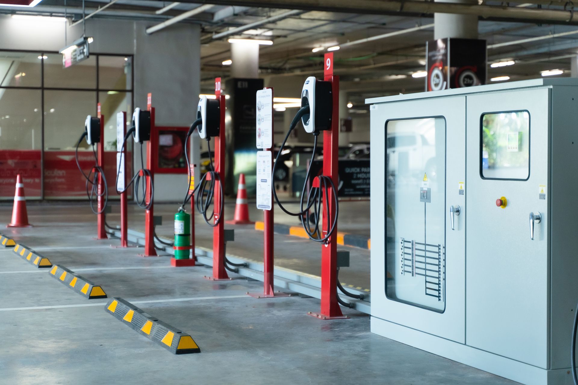 Photo of an indoor electric vehicle charging station. The charging cables are on tall pillars. Nearby raised hazard stripe bumpers restrict the space around the chargers.