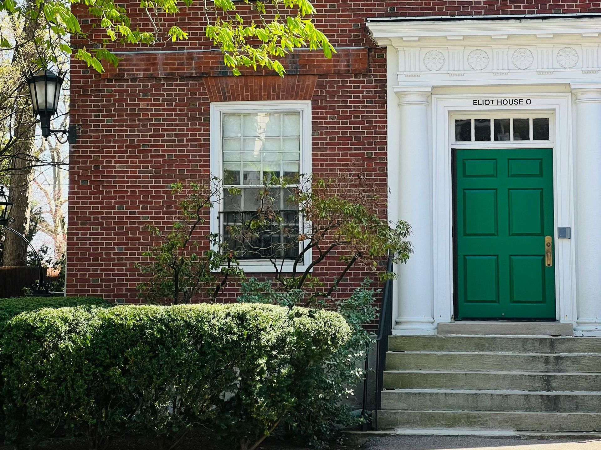 A red-brick building with white-trimmed windows and a bold green door centered at the top of a short flight of stone steps. The doorway is framed by white columns and decorative molding, with the sign
