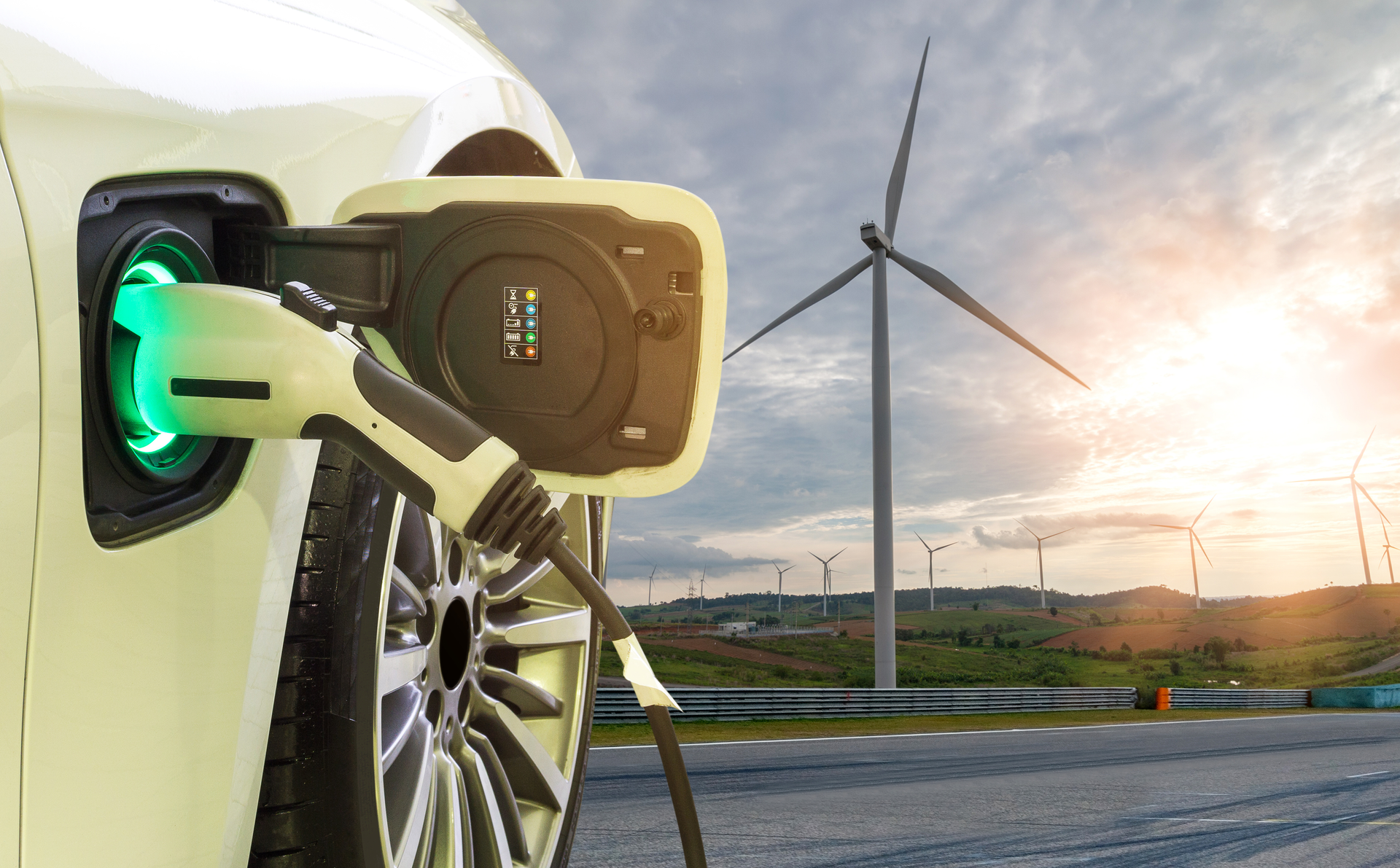 Photo depicting an EV in the foreground, charging though a cable. The car is facing towards a wind farm, lit by the sun.