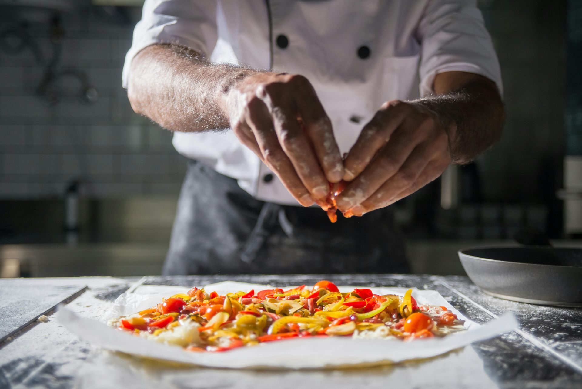Close-up of a chef in a white uniform and black apron preparing a colourful dish on a floured work surface. The chef's hands are in focus, delicately sprinkling ingredients like chopped red and yellow peppers, tomatoes, and onions onto a base, possibly for a pizza or flatbread. The background is softly blurred, suggesting a professional kitchen setting.