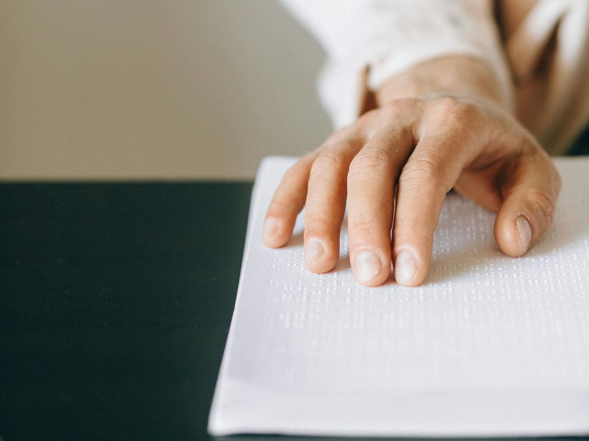 Close-up of a person's hand gently moving across a page of embossed braille text. The fingers are lightly tracing the raised dots, while the other arm rests casually on a table. The scene is softly lit, with a neutral background and a calm, focused atmosphere.