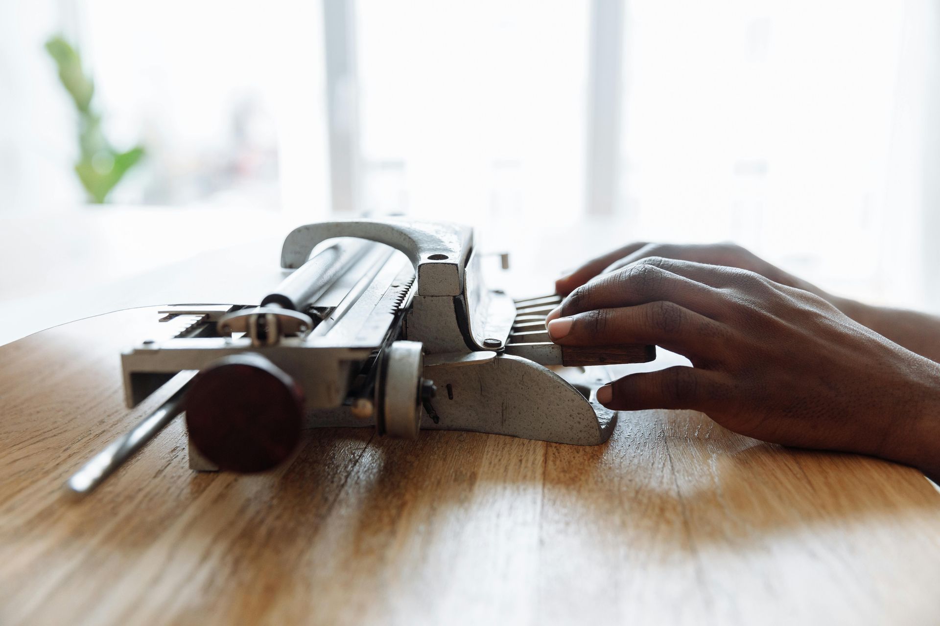 Close-up of a person's hands typing on a mechanical braille writing machine placed on a wooden table. The focus is on the fingers pressing the keys, with soft natural light coming through a large window in the background, creating a calm and focused atmosphere.
