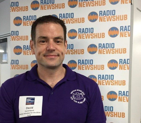 A photograph of David Beard, stood in front of a Radio Newshub banner. He is wearing a British Wireless for the Blind polo shirt with his name on the badge. 