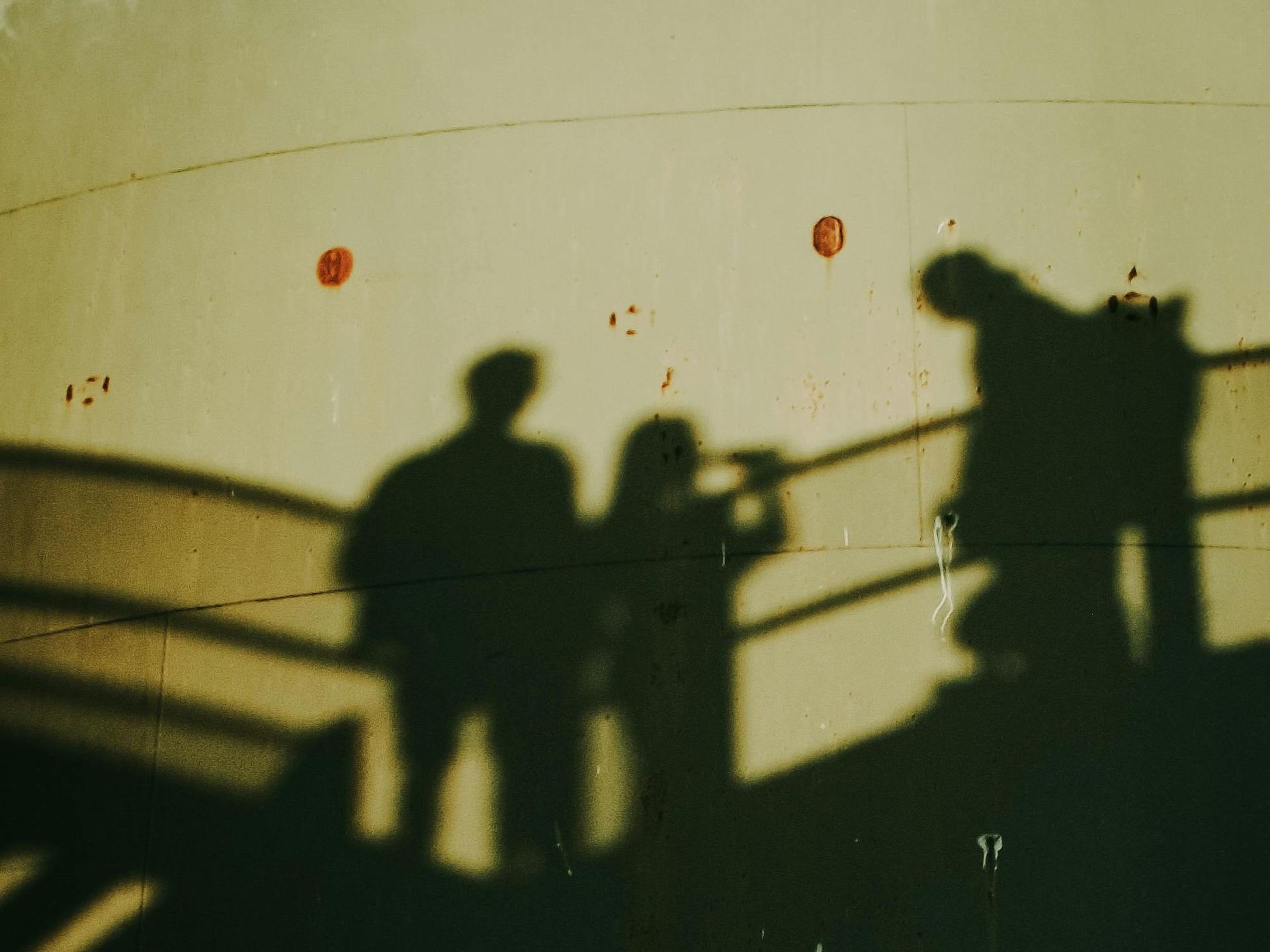 Three human shadows cast sharply onto a large concrete wall, likely by a low sun. The shadows show two figures standing side by side, one appearing to raise a camera or binoculars, and a third figure leaning forward slightly near a railing. Diagonal shadows from metal railings create strong geometric patterns across the surface. The wall is marked by faint rust spots and bolts.