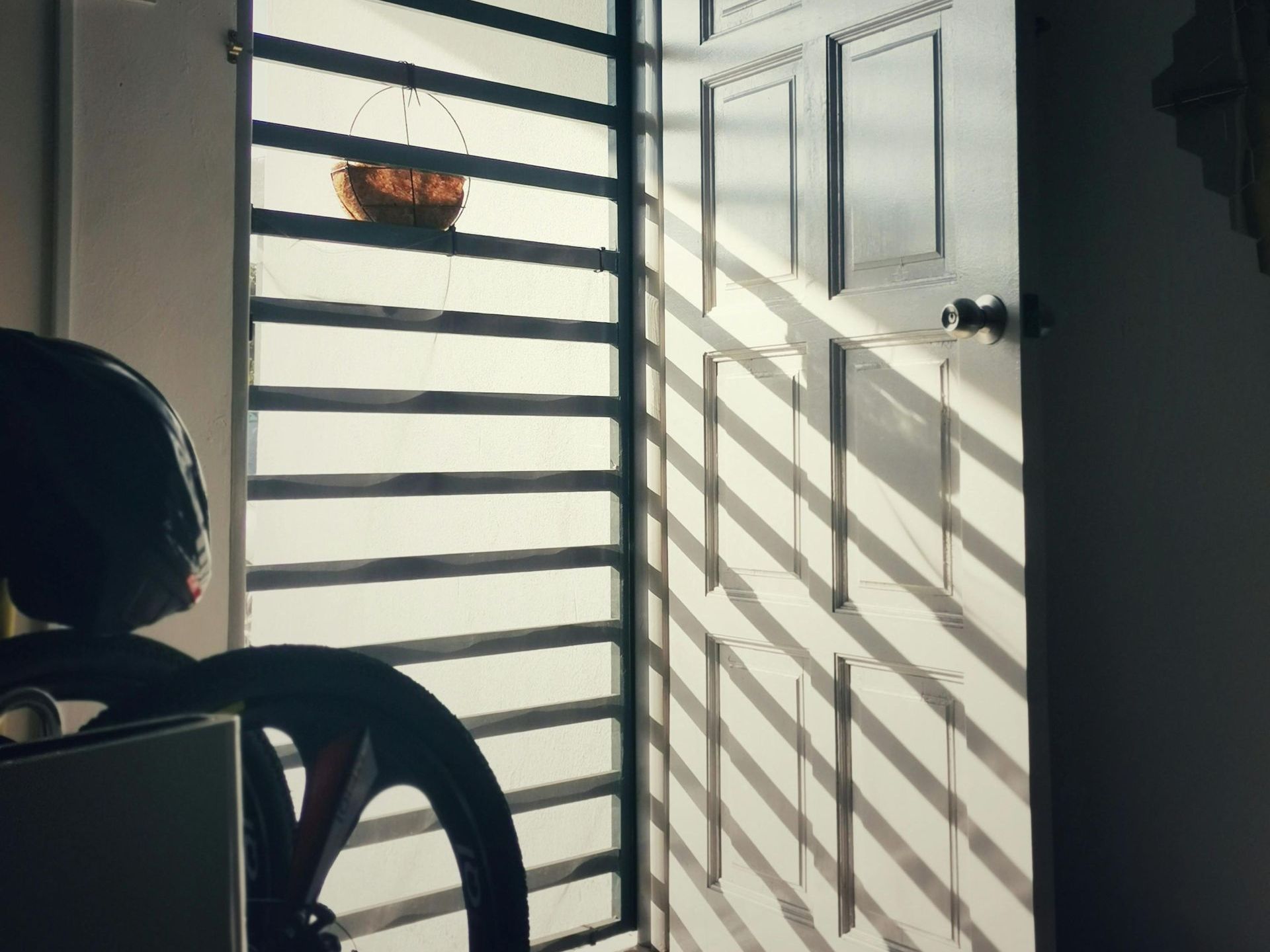 Sunlight streams through a barred security gate and an open wooden door, casting bold diagonal shadows across the tiled floor and door panels. A hanging coconut shell planter is suspended near the bars. In the lower left, part of a bicycle wheel and helmet are partially visible, suggesting an indoor entrance area.