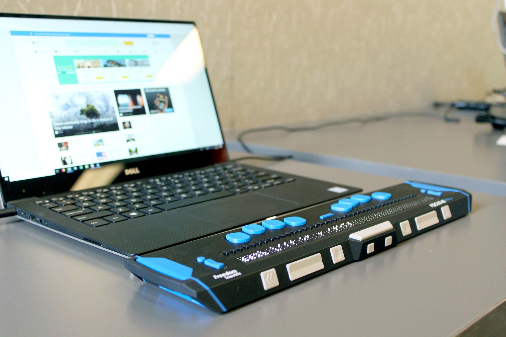 A laptop sits behind a Braille screen reader, its screen displaying a Facebook feed.