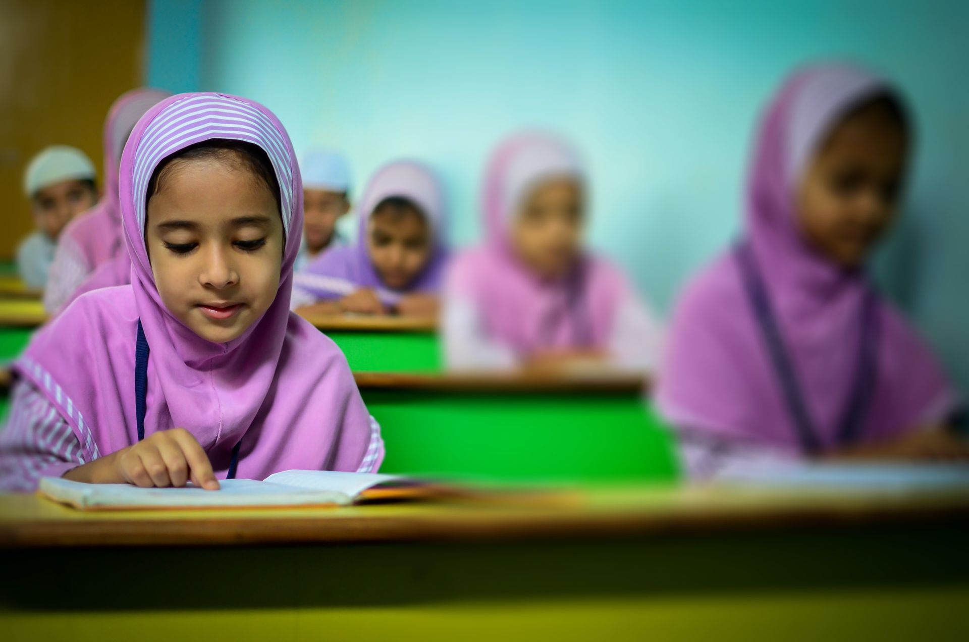 A young girl in a hijab reads from a book, surrounded by other children in a tidy classroom.