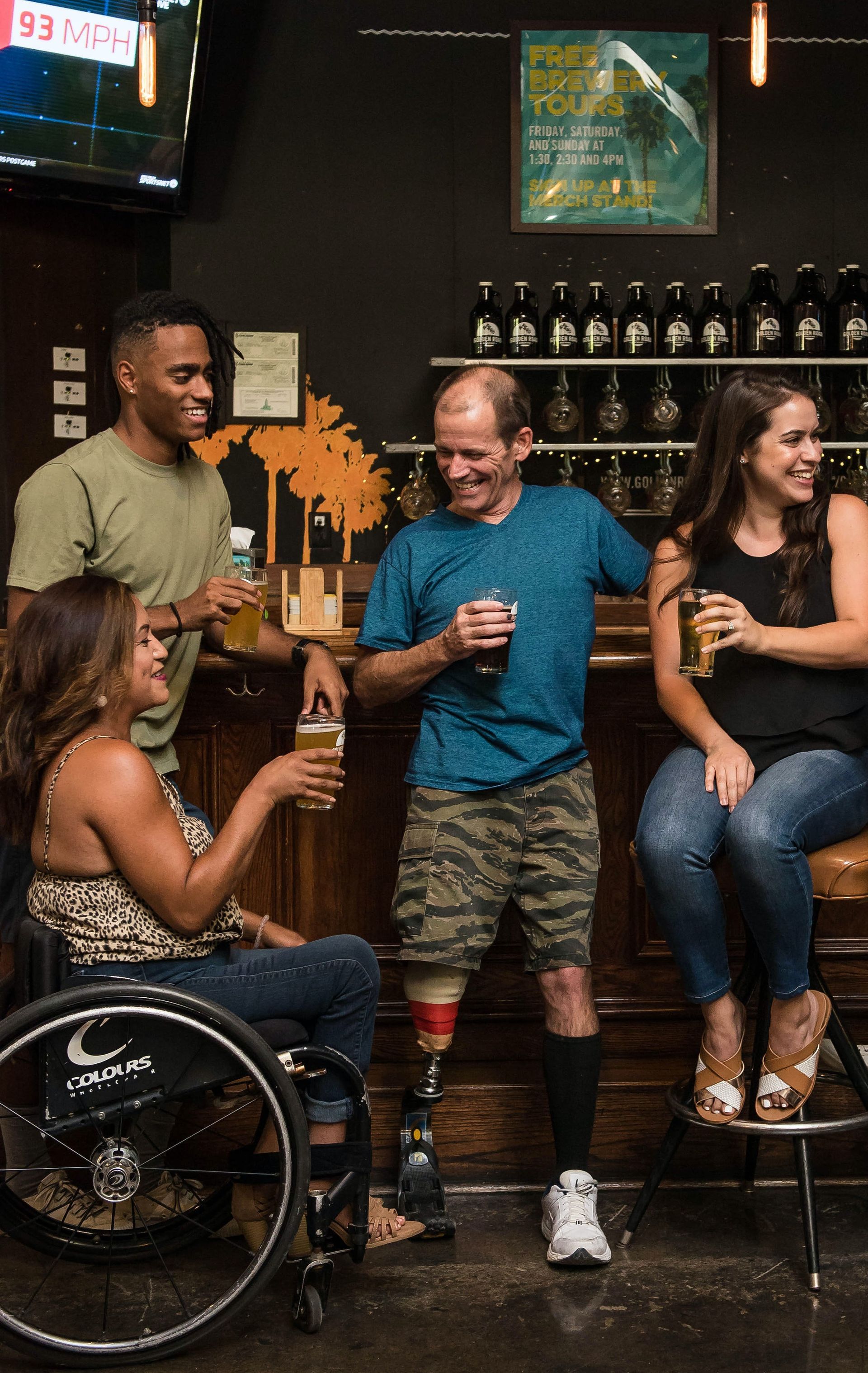 A group of four friends share a drink in a pub, one of the two women in a wheelchair and one of the two men with an amputated leg.