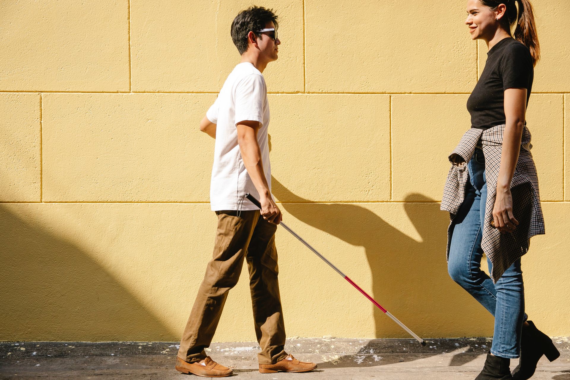(Outdoor Adventures) A young man with sight loss walks across a street, using his white cane to guide him.