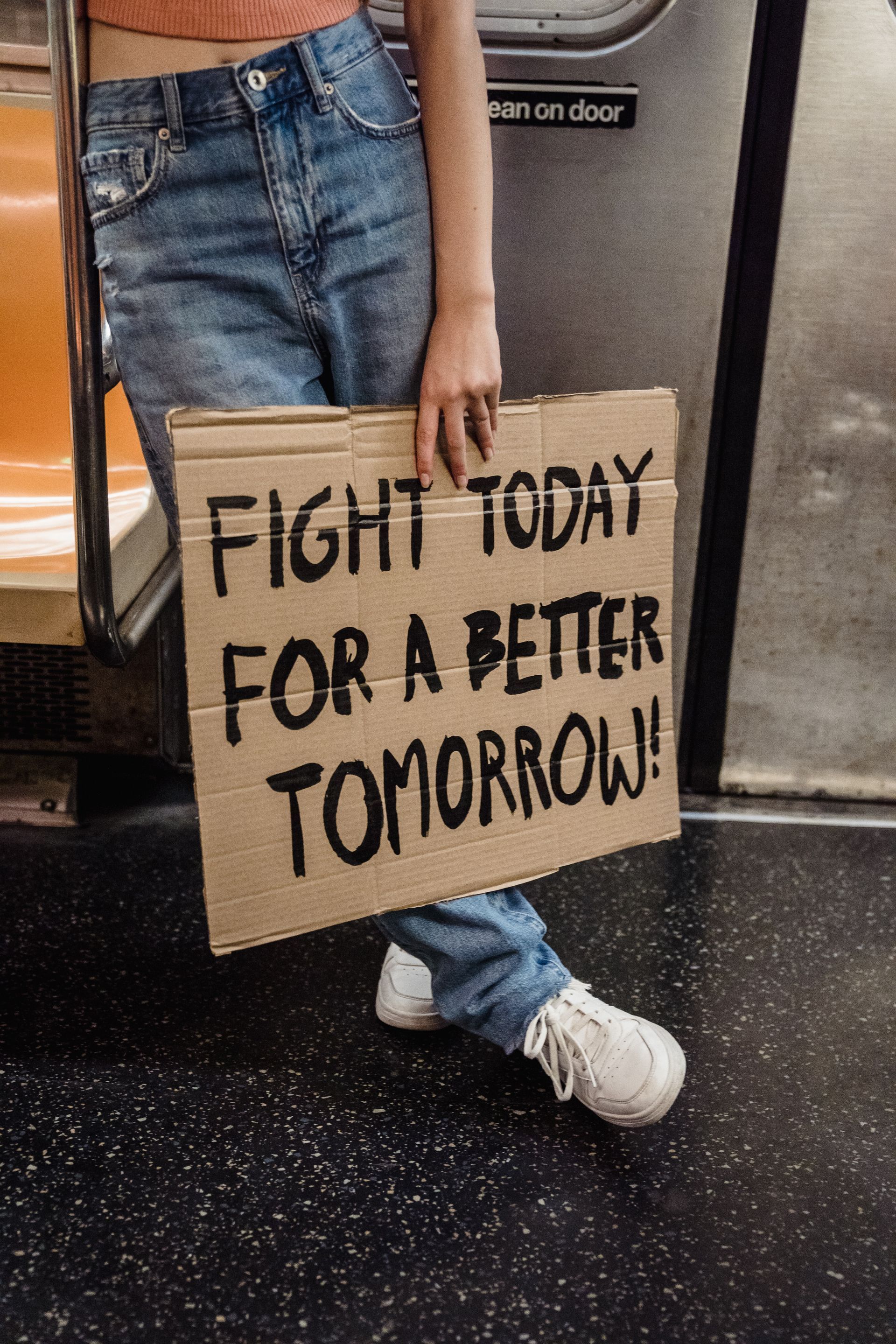 A young woman waits on a train, holding a sign which declares 'Fight today for a better tomorrow.'