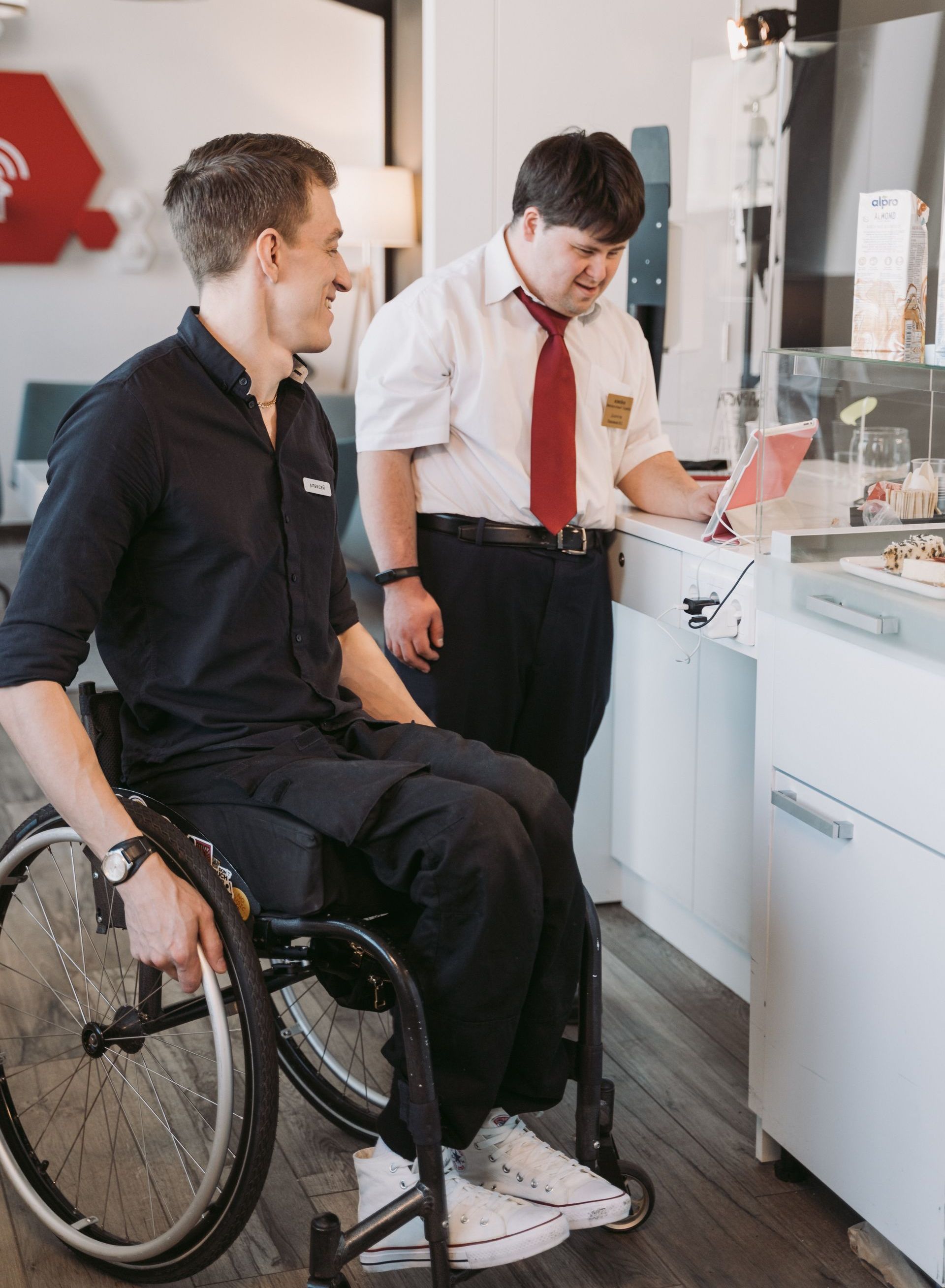 A male supervisor with Down Syndrome and a cafe worker in a wheelchair looking on a computer behind the counter.