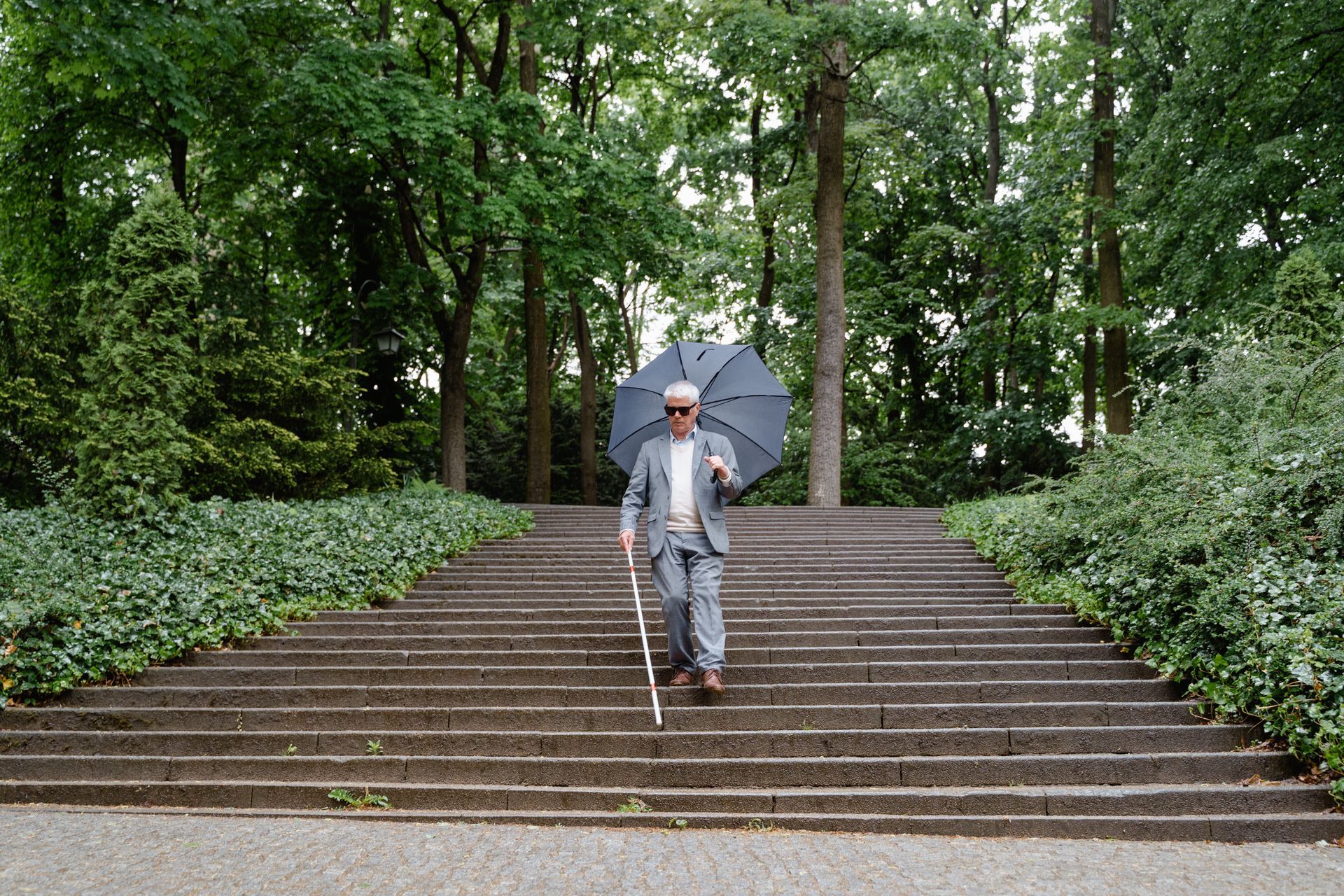 An older, smartly dressed gentleman holding an umbrella navigates down stone steps using a white cane.