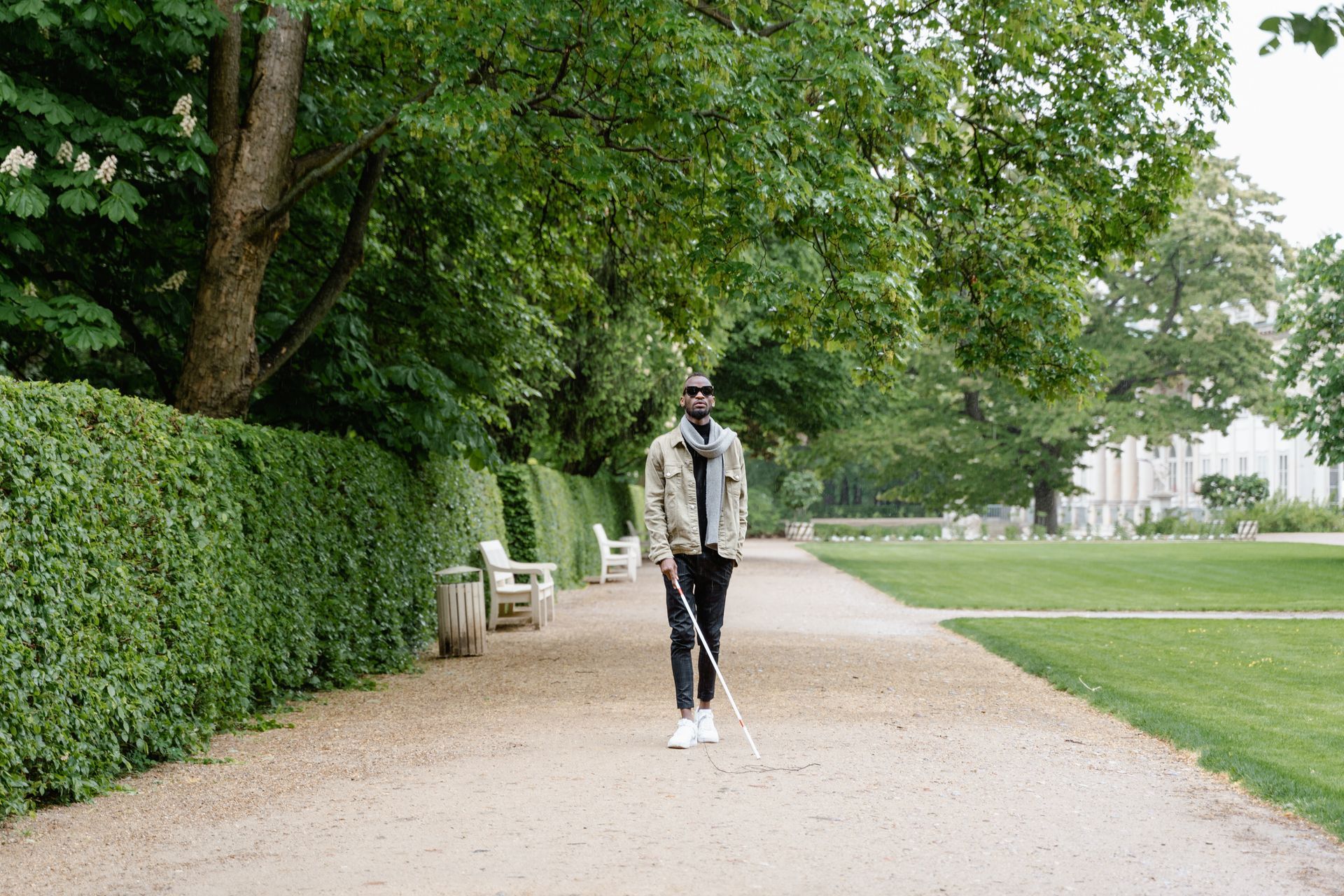 A young man with sight loss strolls in a heavily manicured park, using his white cane to locate obstacles.