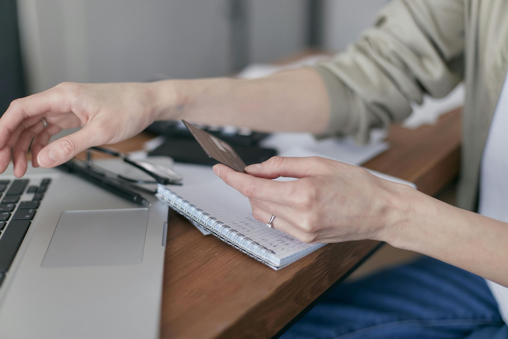 Photograph of a person sat at a desk holding a bank card. With their other hand they reach towards a laptop.