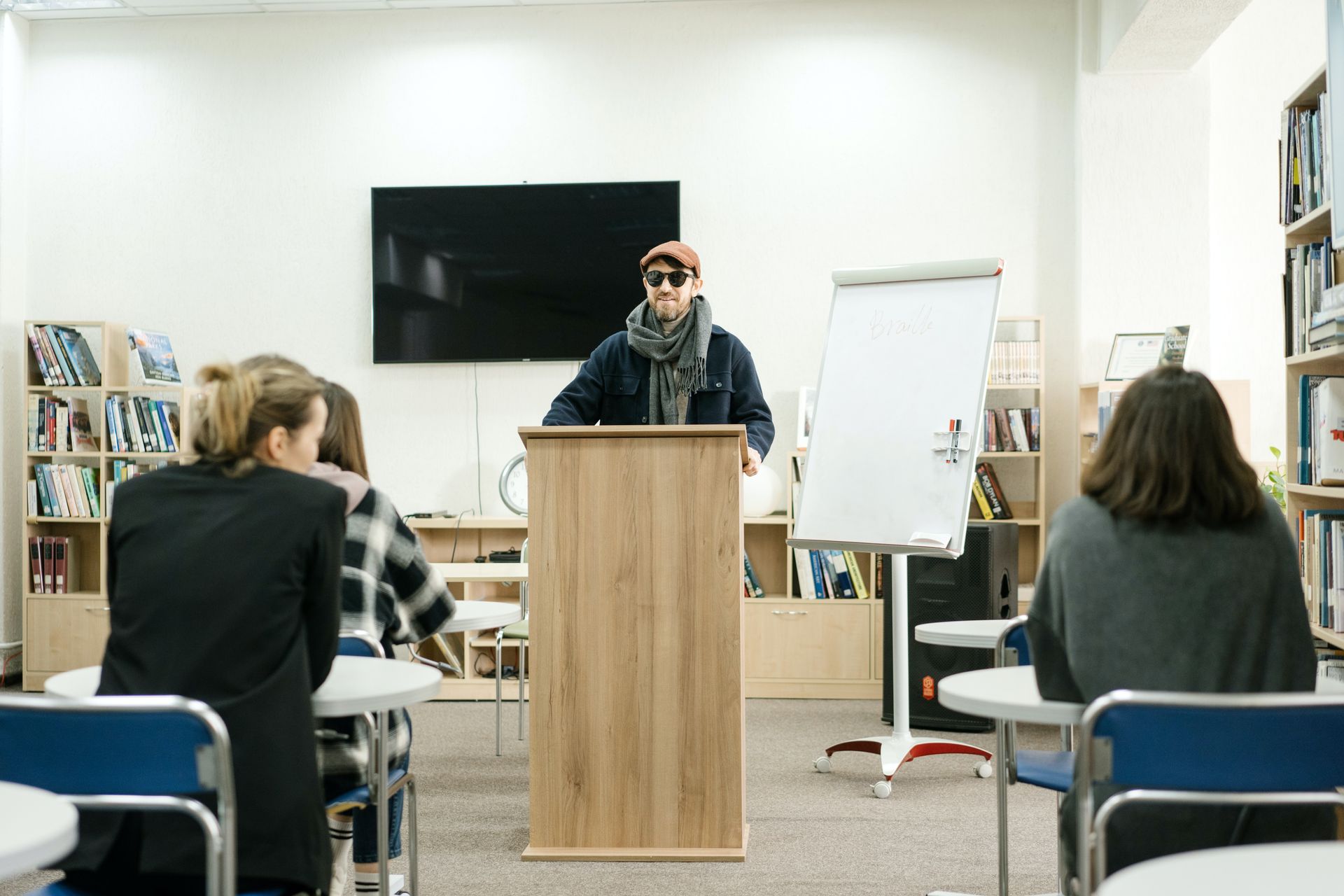 A man with sight loss stands at a lecturn, conducting a speech in front of a small class of mature learners.