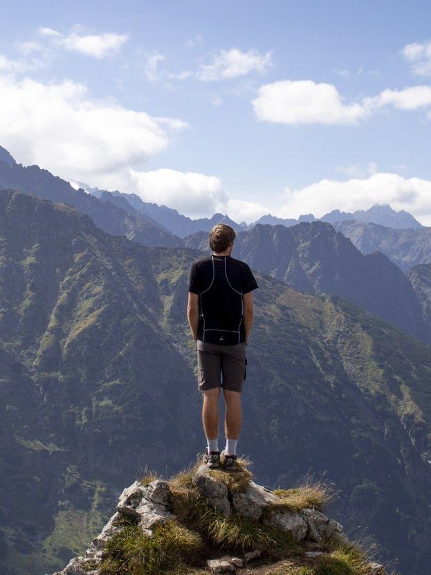 A man stood atop a grass-topped peak, gazing towards mountains crowned by clouds.