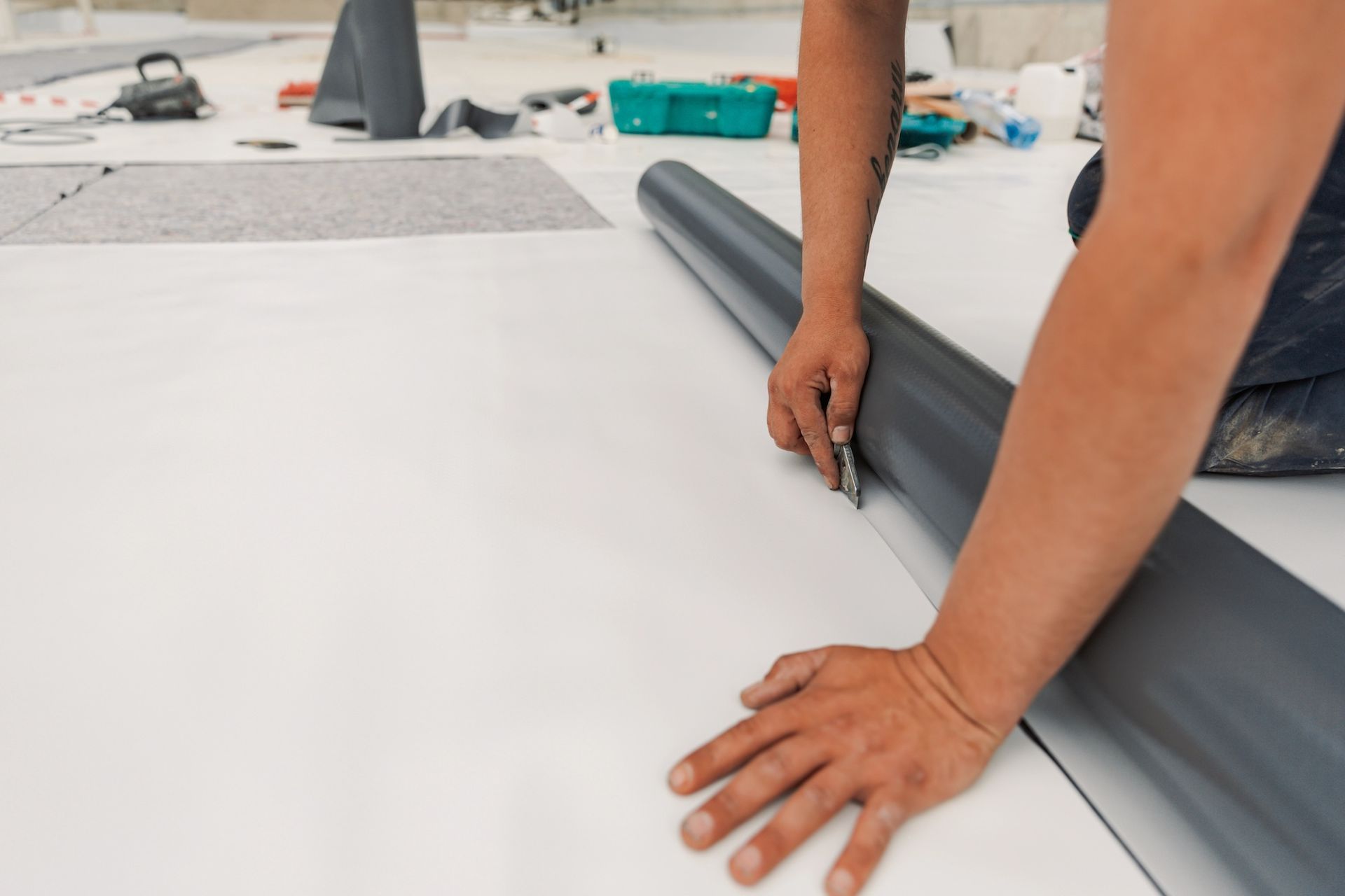 A person cutting rolled white flooring with a utility knife, tools and materials in the background.